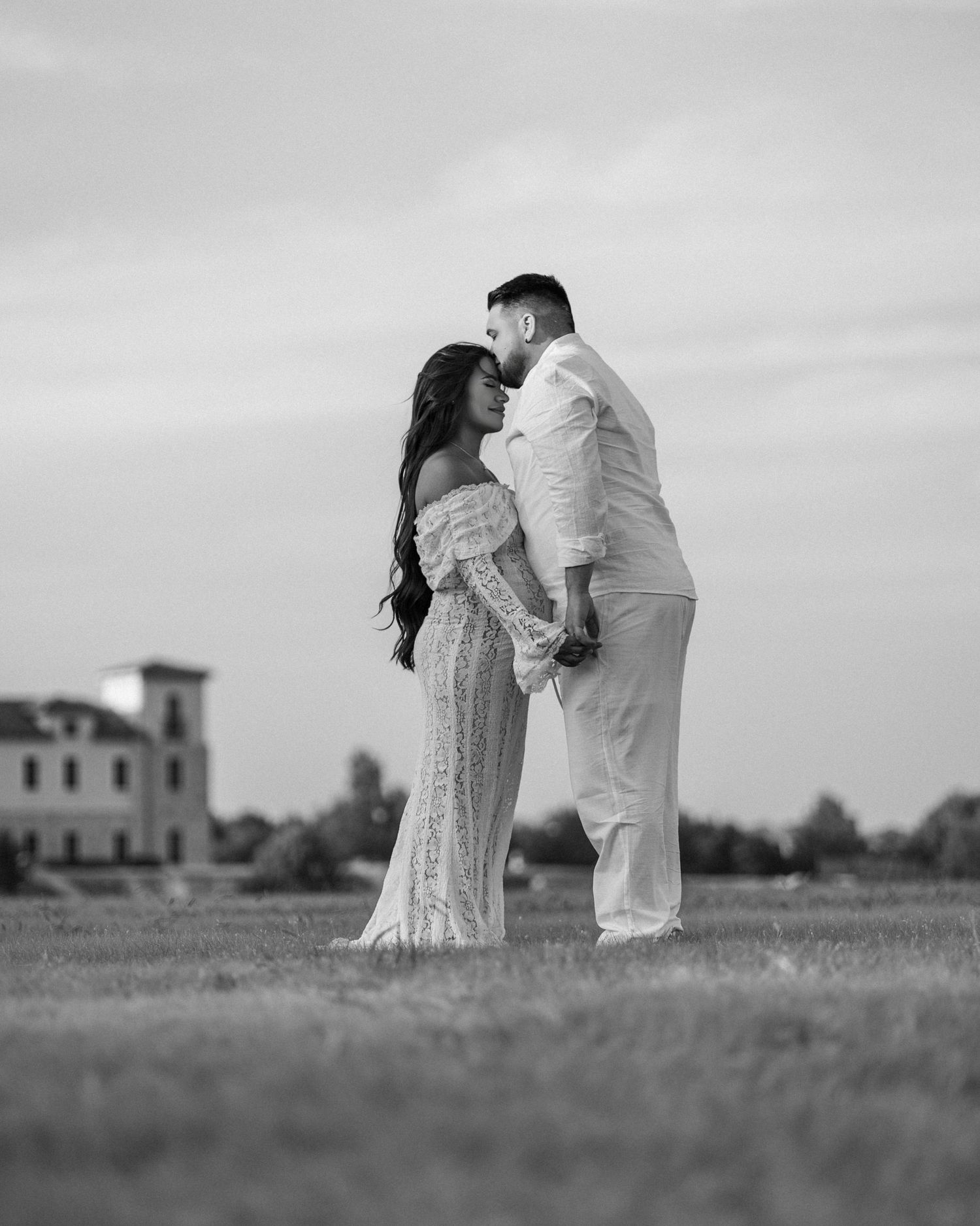 Romantic couple kneeling in a grassy field with a building in the distance.