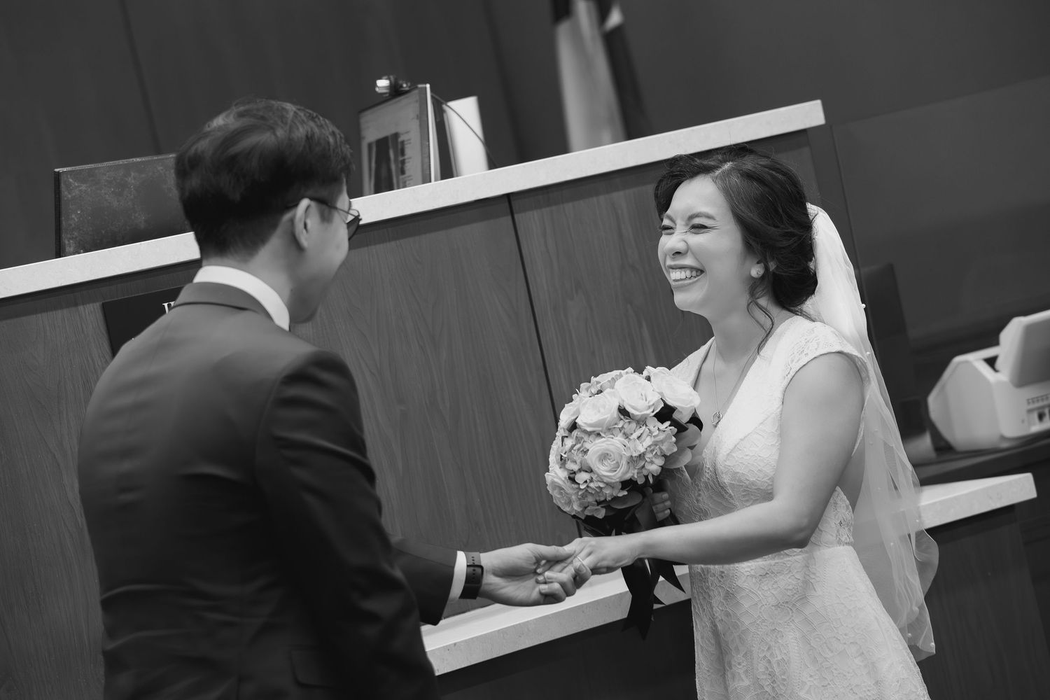 Black and white photo of a smiling bride holding bouquet exchanging vows with groom at wedding ceremony.