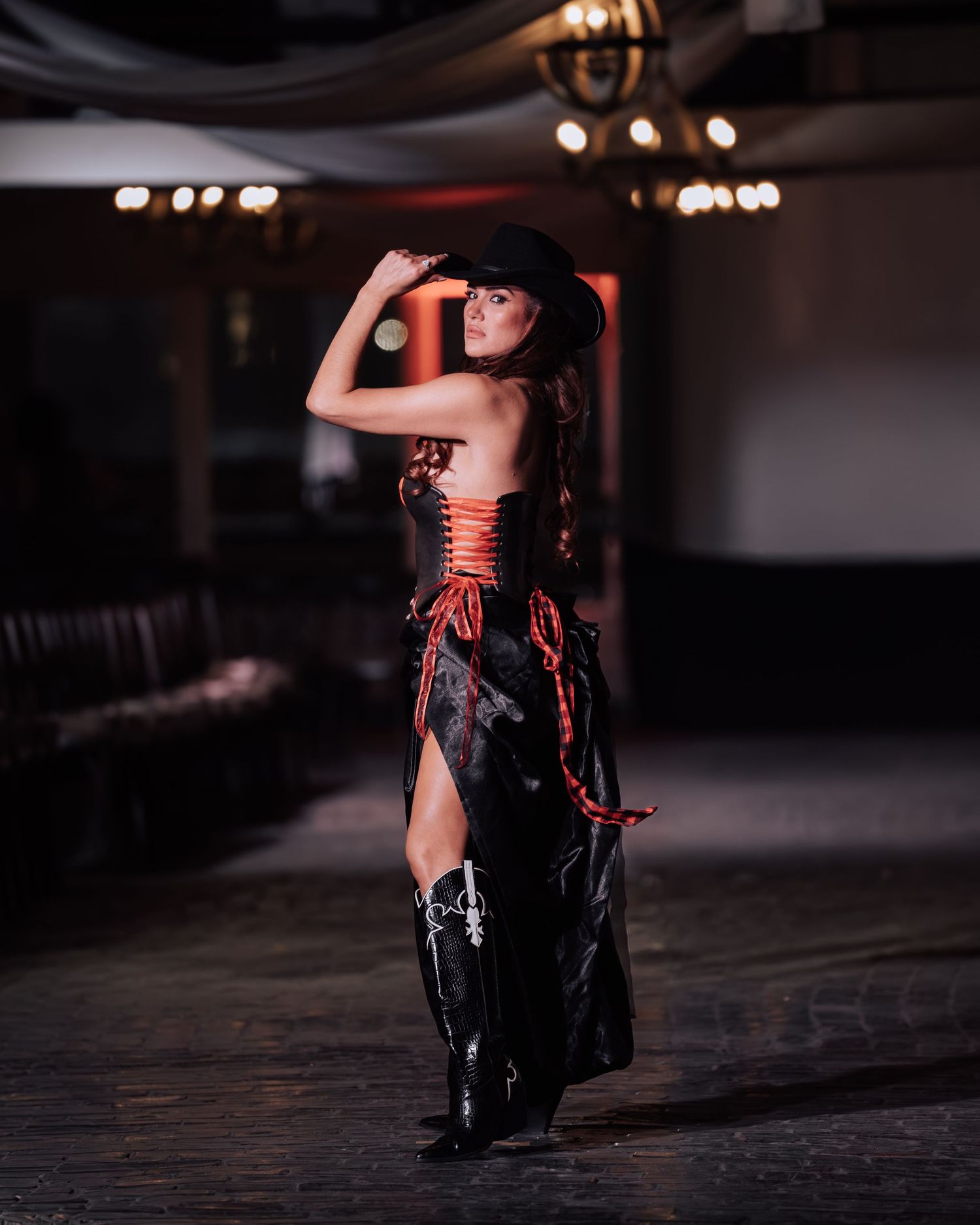A dramatic fashion shoot featuring a model in black latex and red corset posing in a dimly lit industrial space.