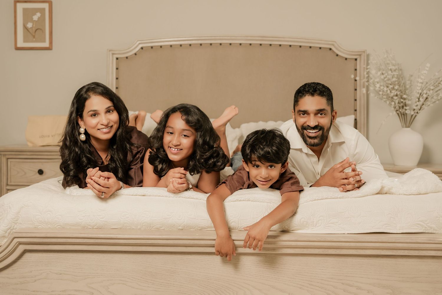 A happy South Asian family of four lying on a white bed, smiling together in a bright, cozy bedroom.
