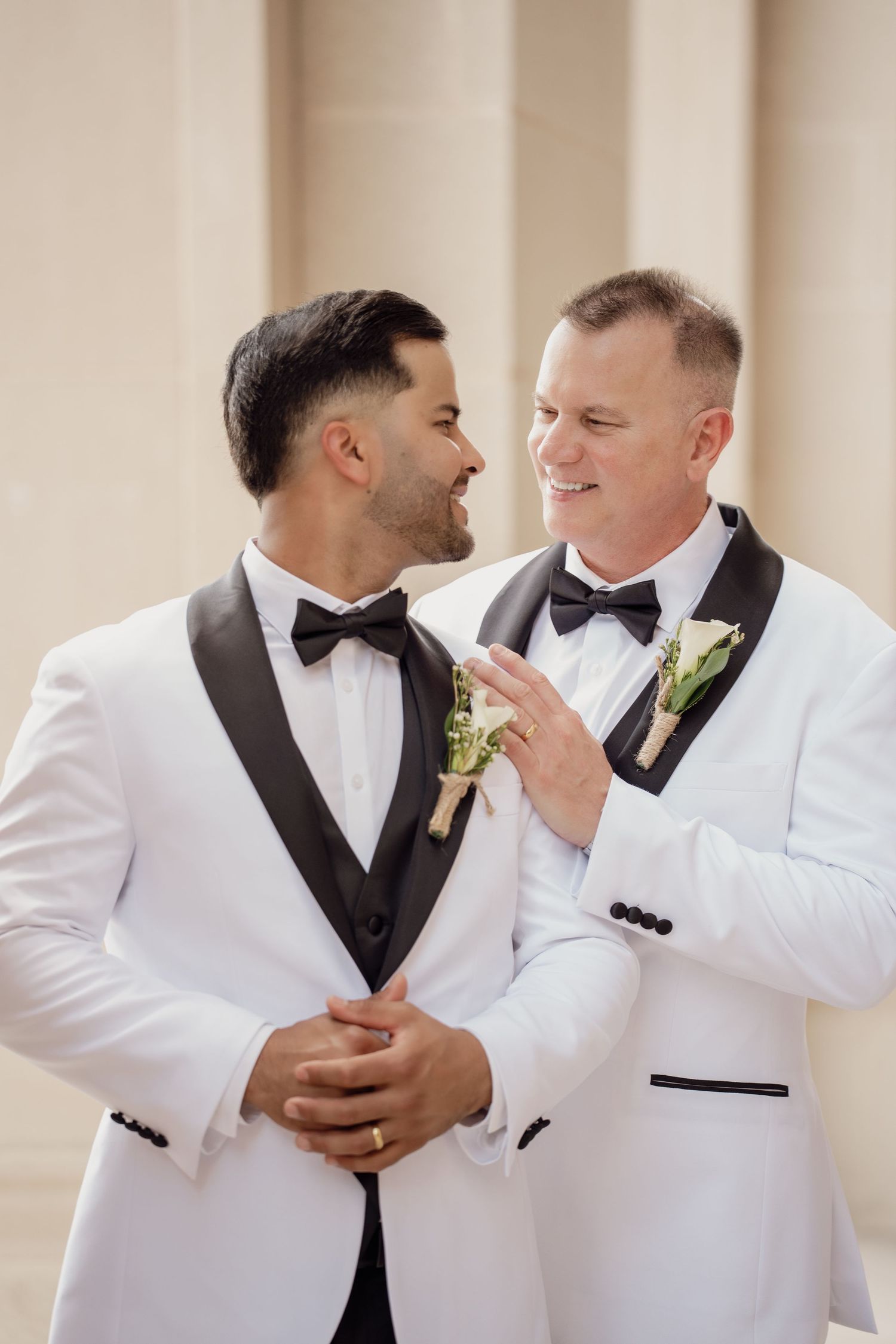 Two grooms in matching white tuxedos with black lapels smile at each other, wearing boutonnieres on their wedding day.