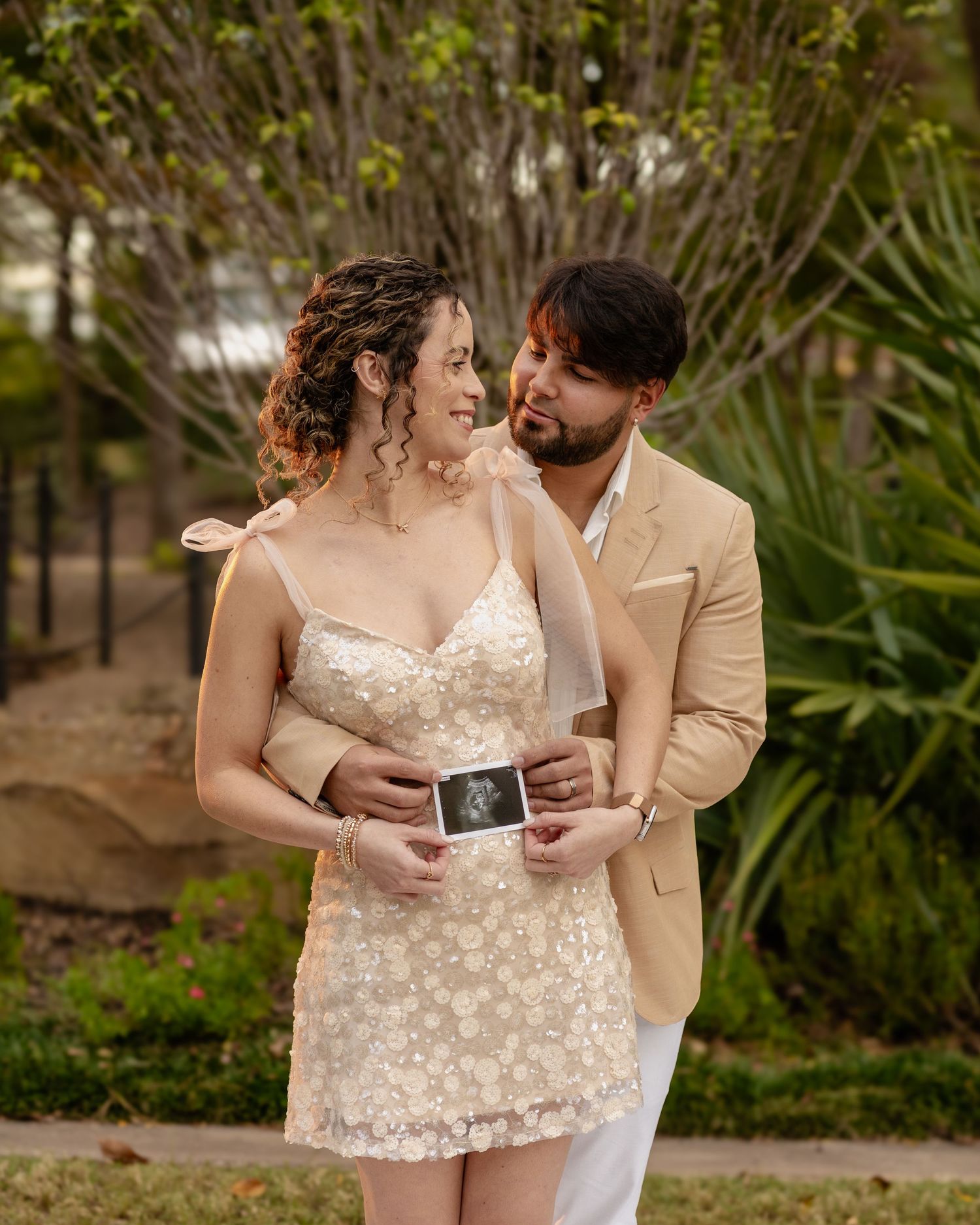 Couple in beige and floral dress sharing intimate moments while holding photos in garden setting.