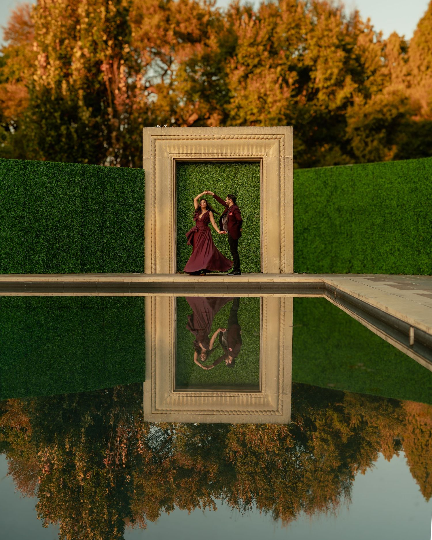 Elegant couple framed by architectural elements with autumn trees reflected in a pool.