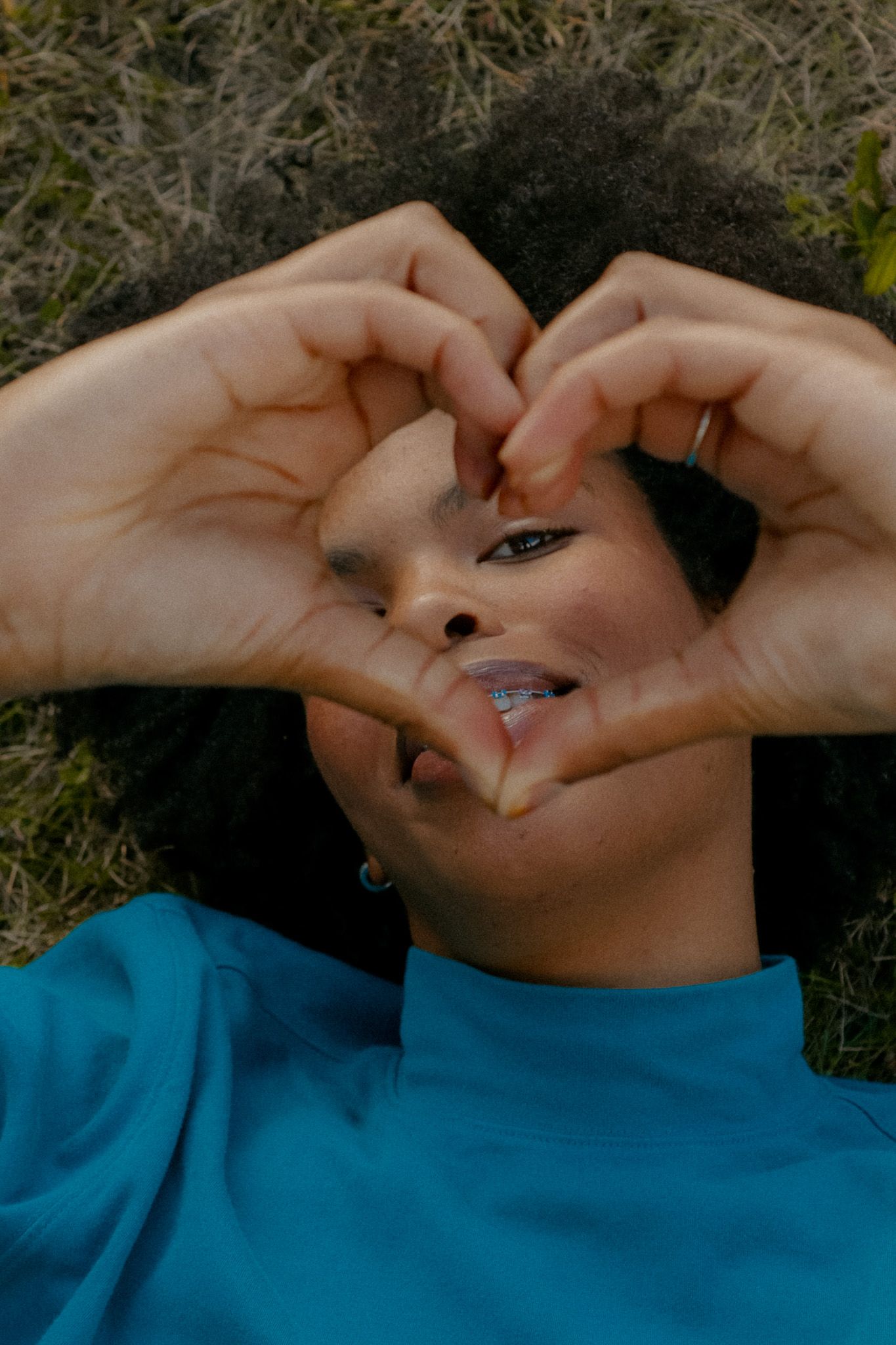 A young woman lying on the grass, making a heart shape with her hands over her face, wearing a blue sweatshirt.