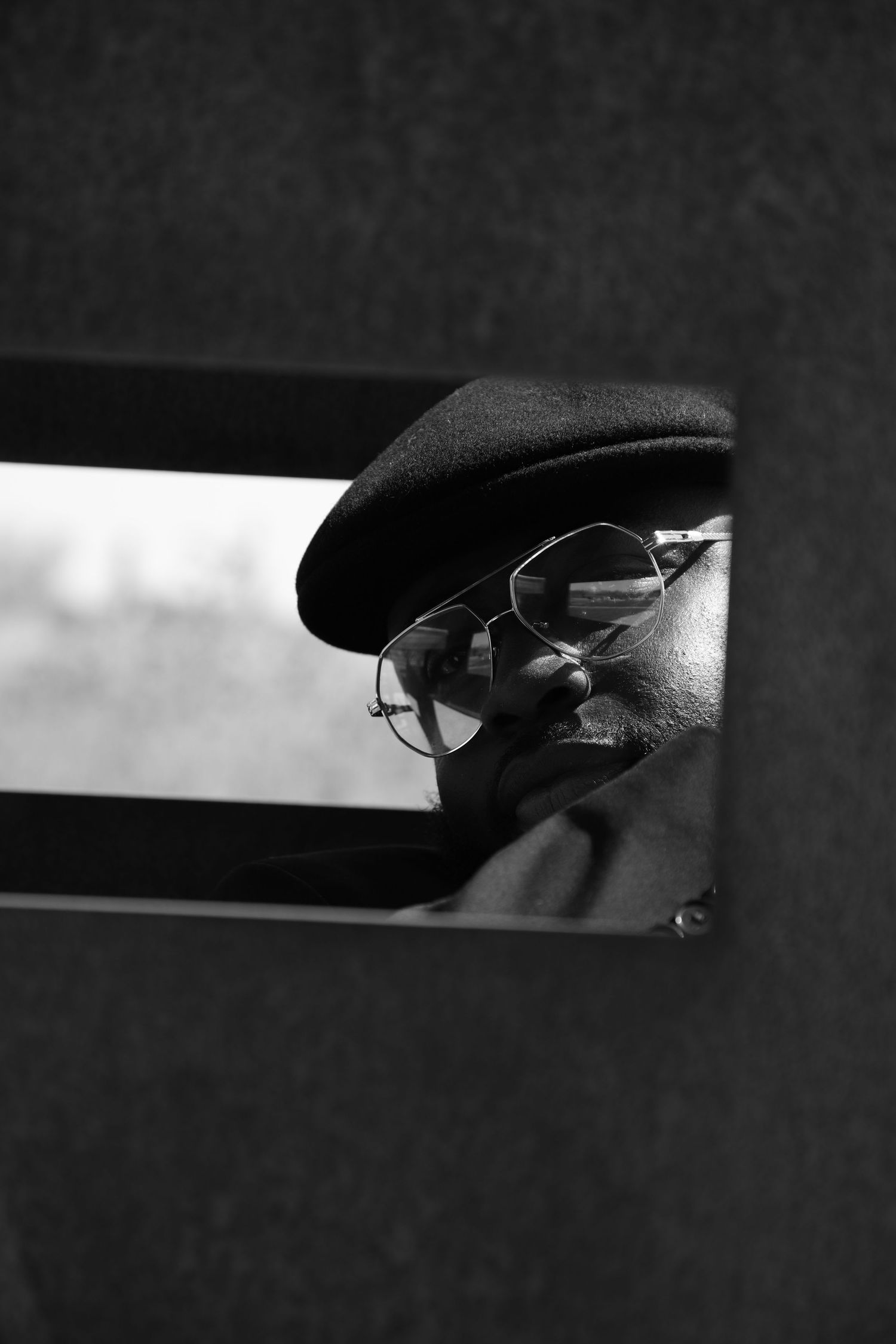 Black and white photo of a person wearing a beret and aviator glasses, partially visible through a small window.