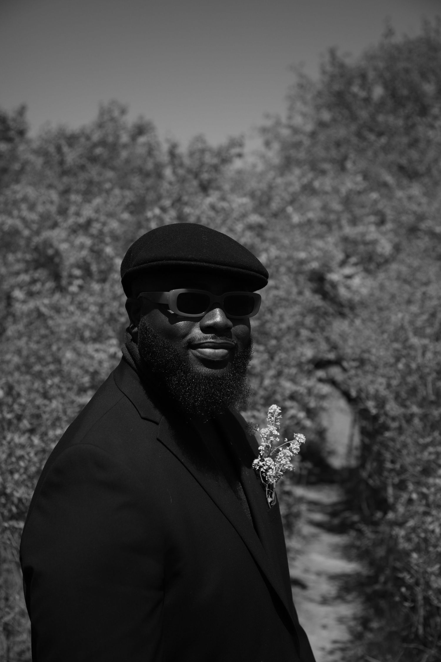 Bearded Black man in black cap, sunglasses, and blazer with floral pin smiles outdoors in black and white photo.