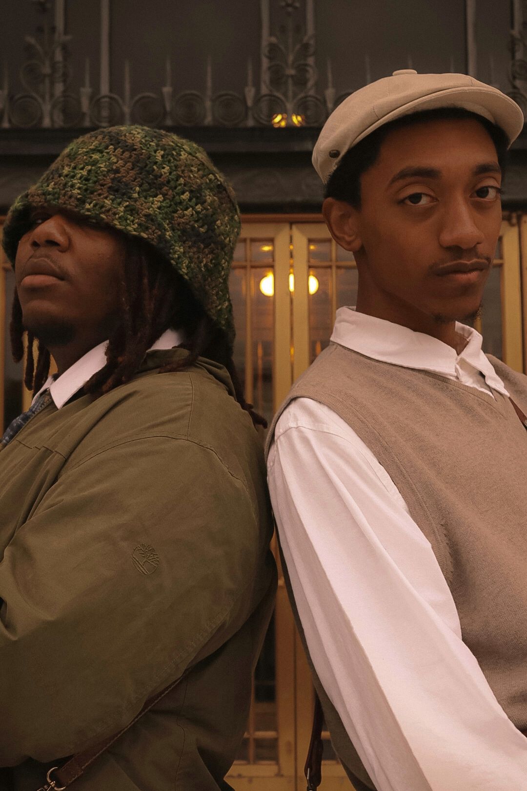 Two young Black men in vintage-style outfits stand back to back posing seriously indoors.