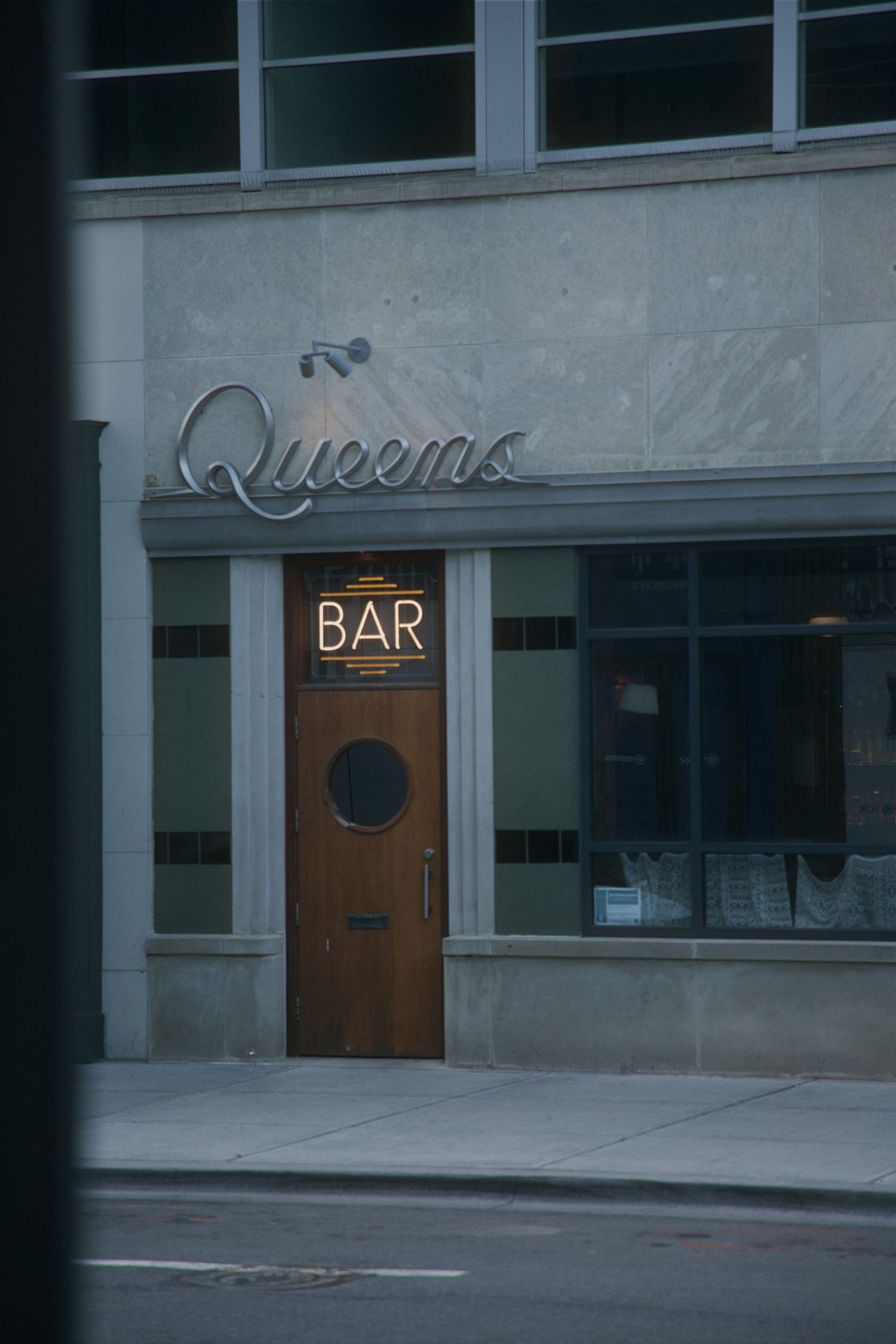 Exterior of Queens Bar featuring a glowing neon sign and wooden door with a porthole window on a city street. Downtown Detroit.