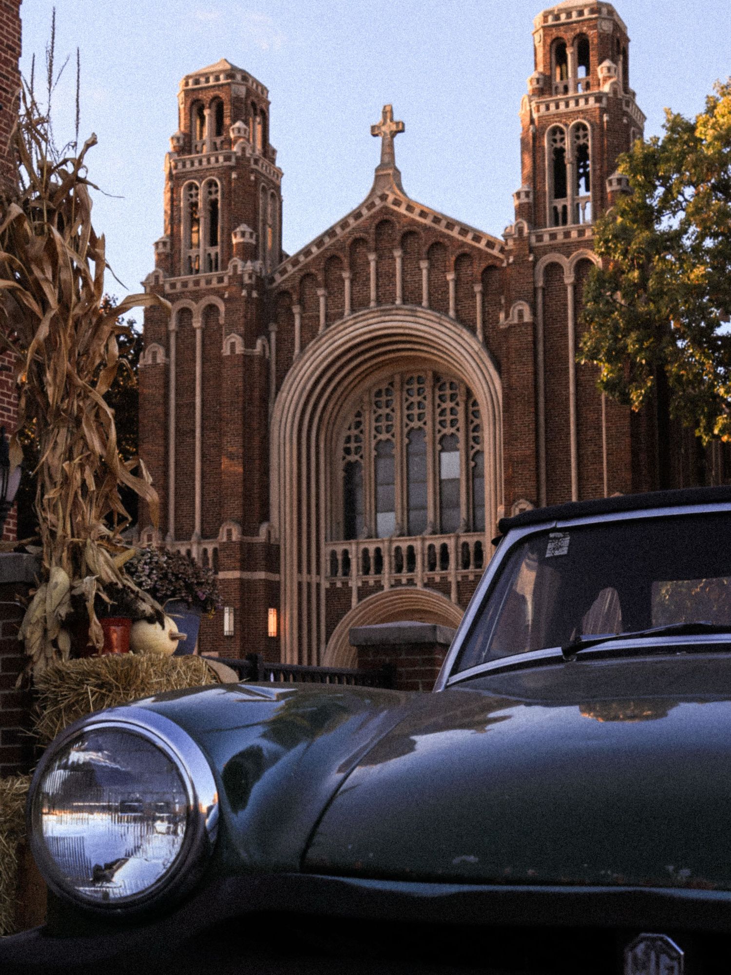 Vintage green MG car parked in front of a Gothic brick church with twin towers and a cross, surrounded by autumn corn stalks.