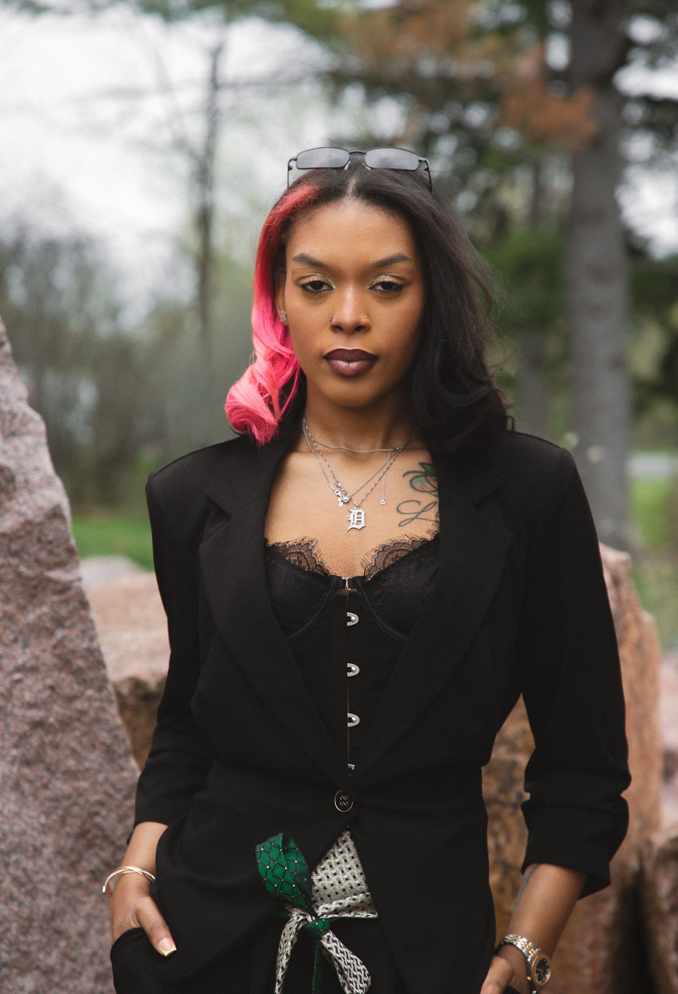 Stylish woman in black outfit with pink hair highlights and tattoos posing outdoors near rocks.
