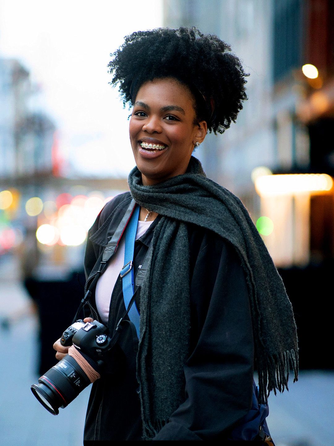 Smiling Black woman photographer holding a DSLR camera outdoors on a city street wearing a gray scarf.