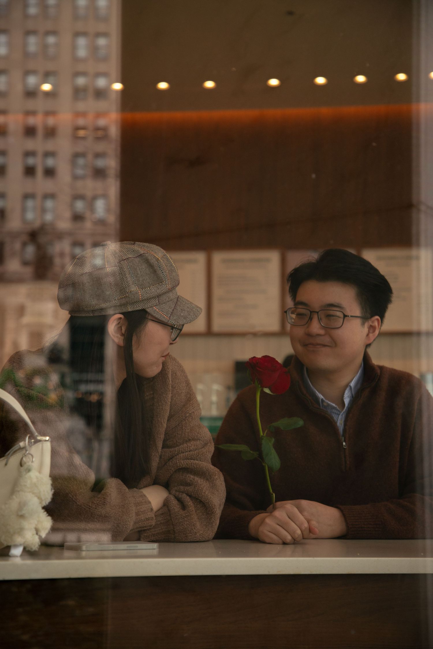 Couple on a romantic date in a cafe, man holding a red rose, viewed through a window.