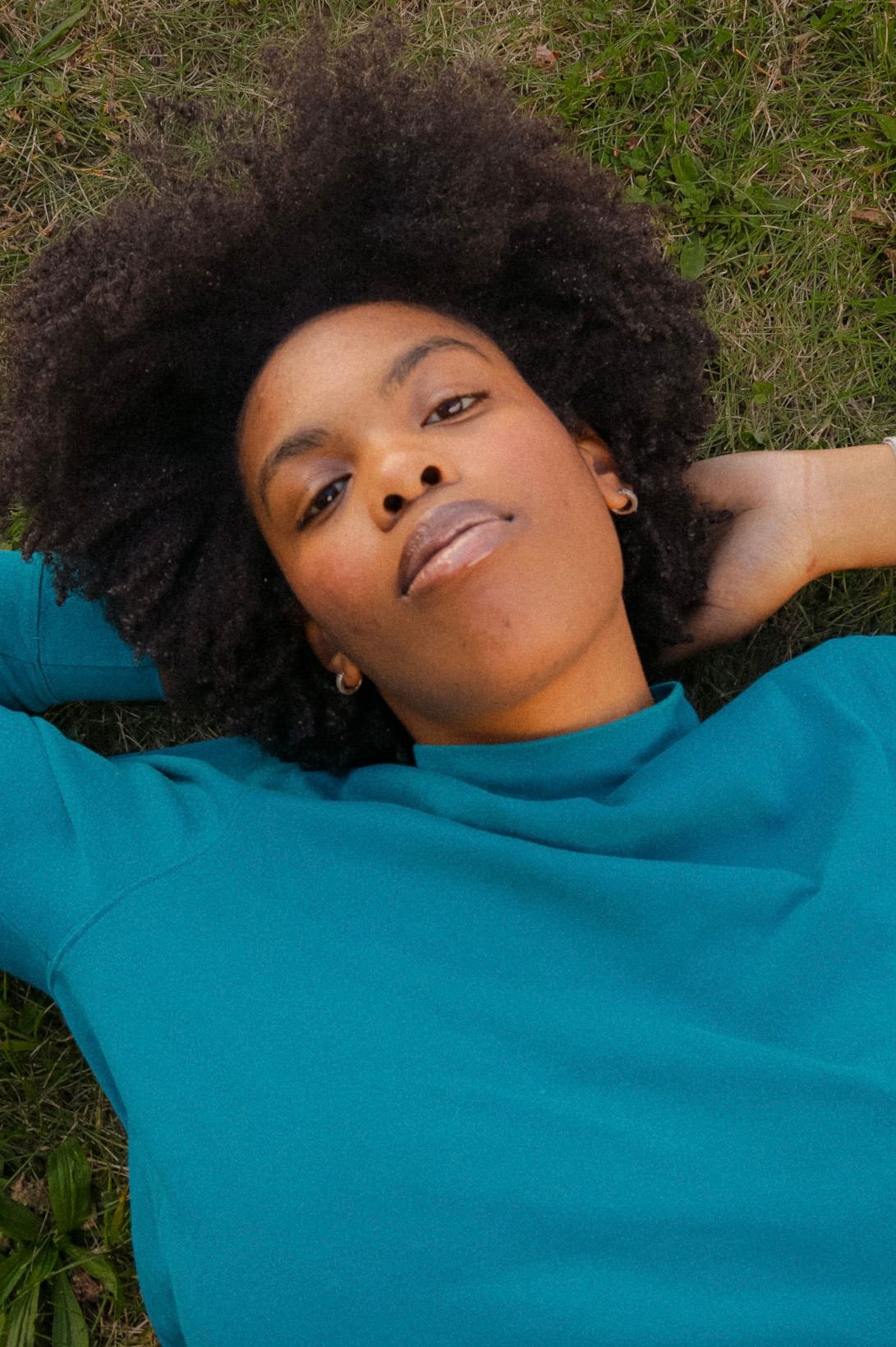 Young Black woman with natural afro hair lying on grass, wearing a teal sweatshirt, looking up peacefully.