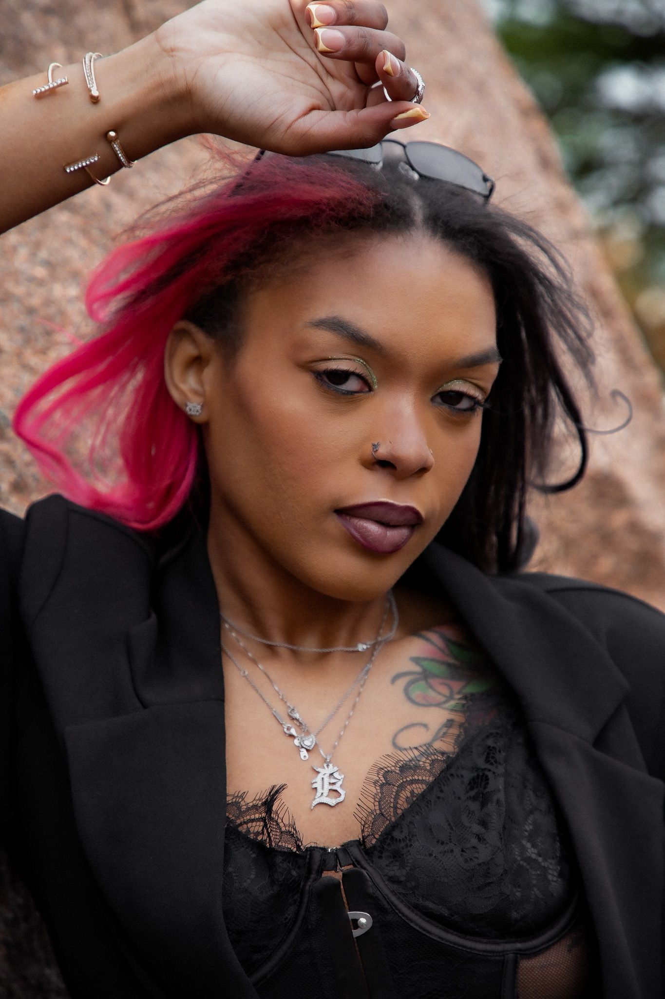 A Black woman with pink and black hair poses outdoors wearing a black blazer, lace top, and silver necklaces.