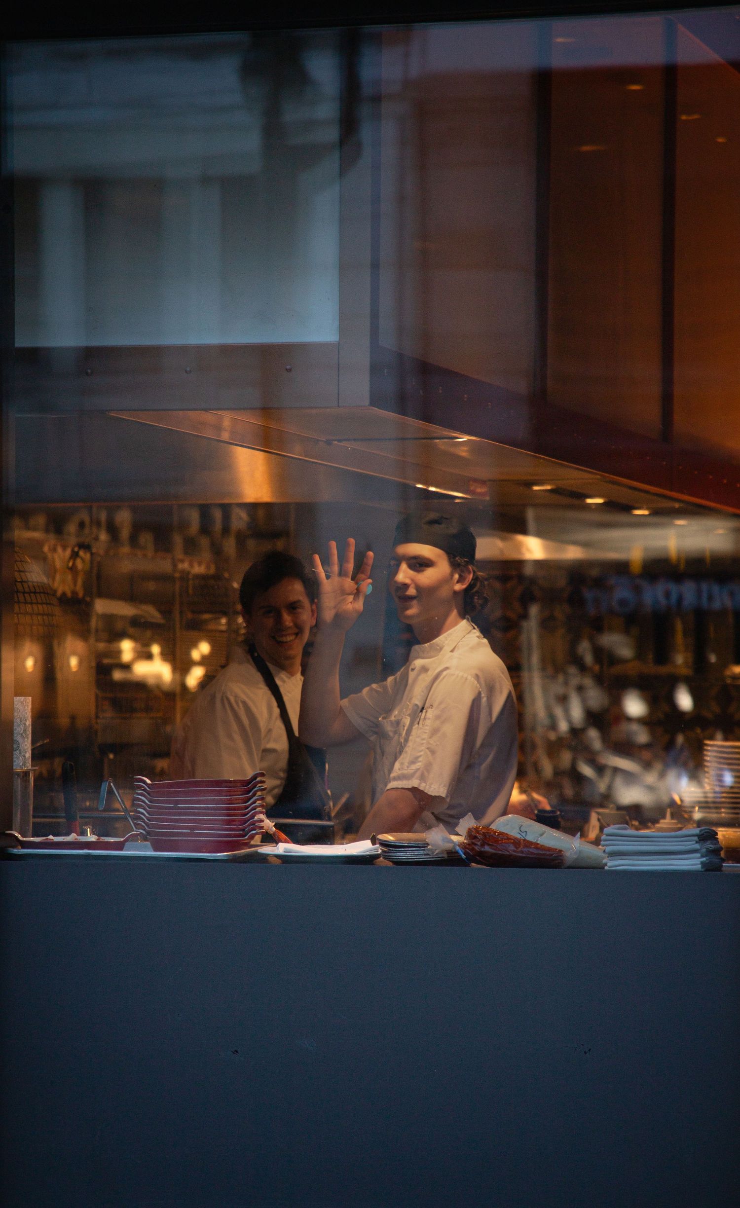 Two restaurant workers smile through a kitchen window, one giving an OK hand gesture near stacked plates.
