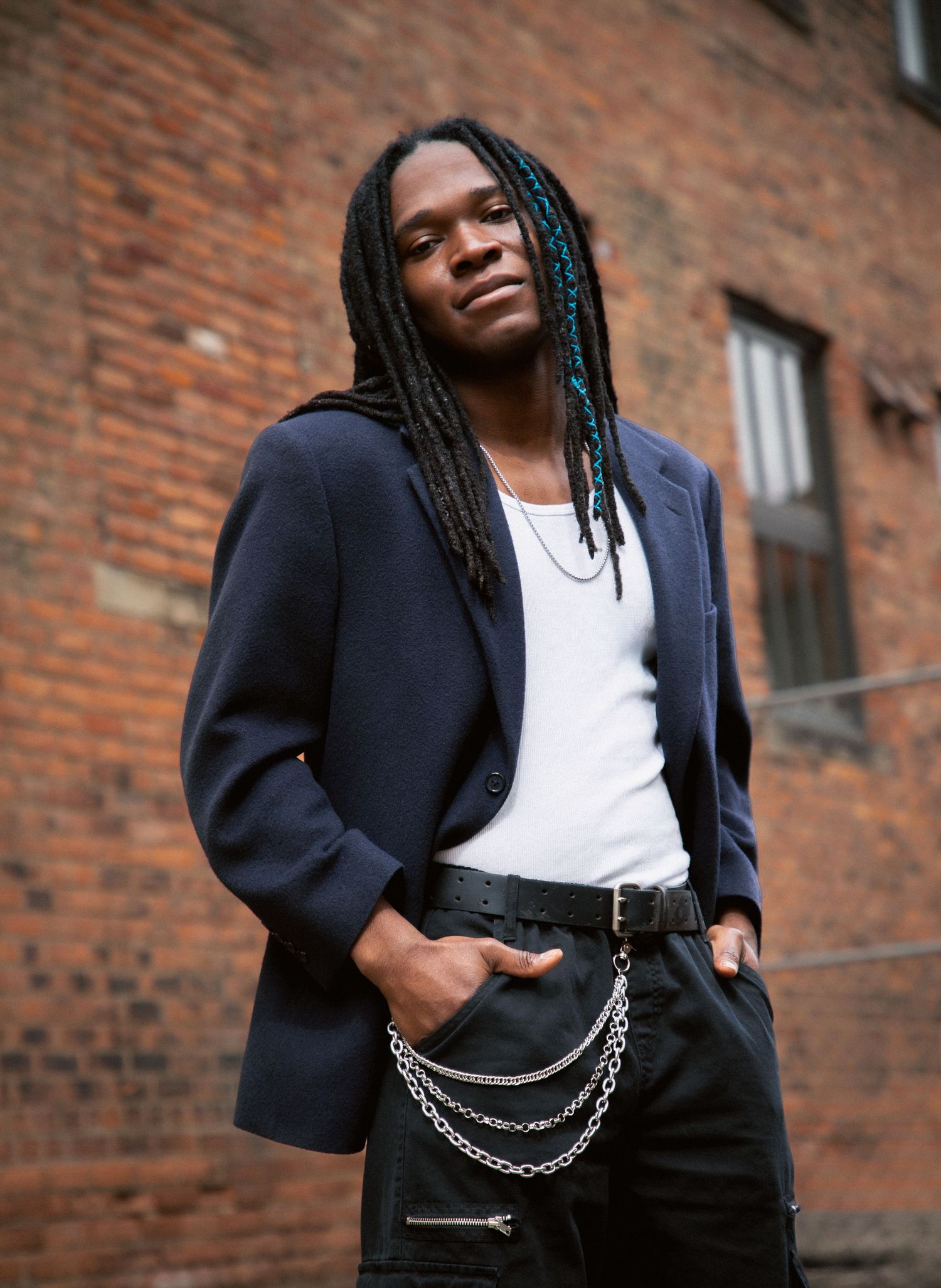 Young man with long braids wearing a navy blazer and chain belt poses confidently against a brick wall.