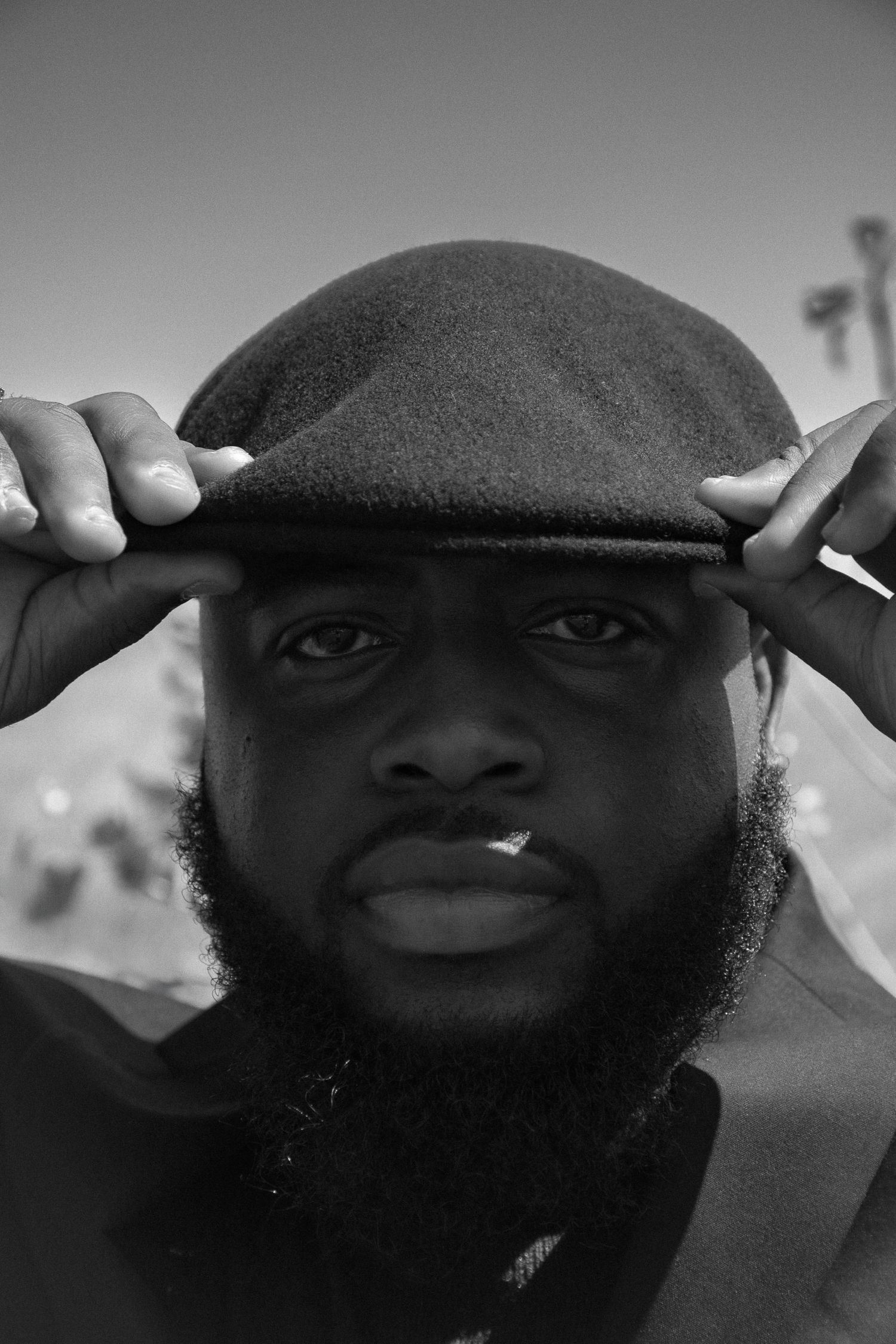 Black and white portrait of a bearded man adjusting a flat cap hat, looking directly at camera.