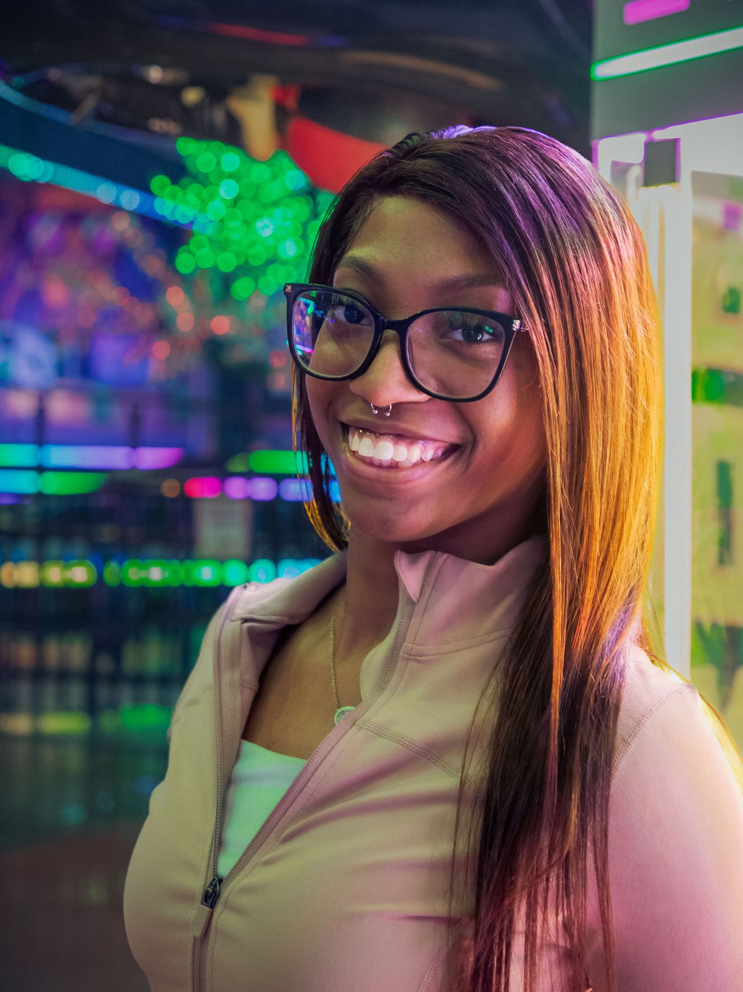 Smiling young woman with glasses and nose ring poses in colorful neon-lit arcade venue.