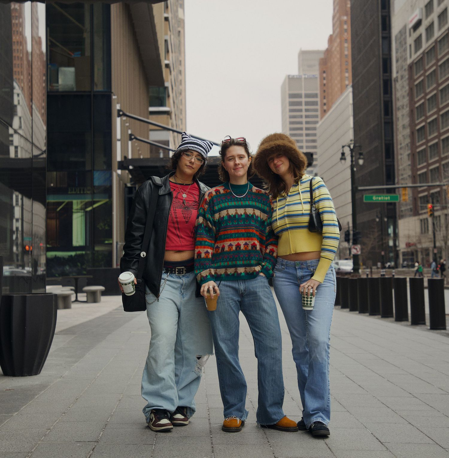 Three stylish friends pose together on a city sidewalk wearing colorful vintage sweaters and jeans.