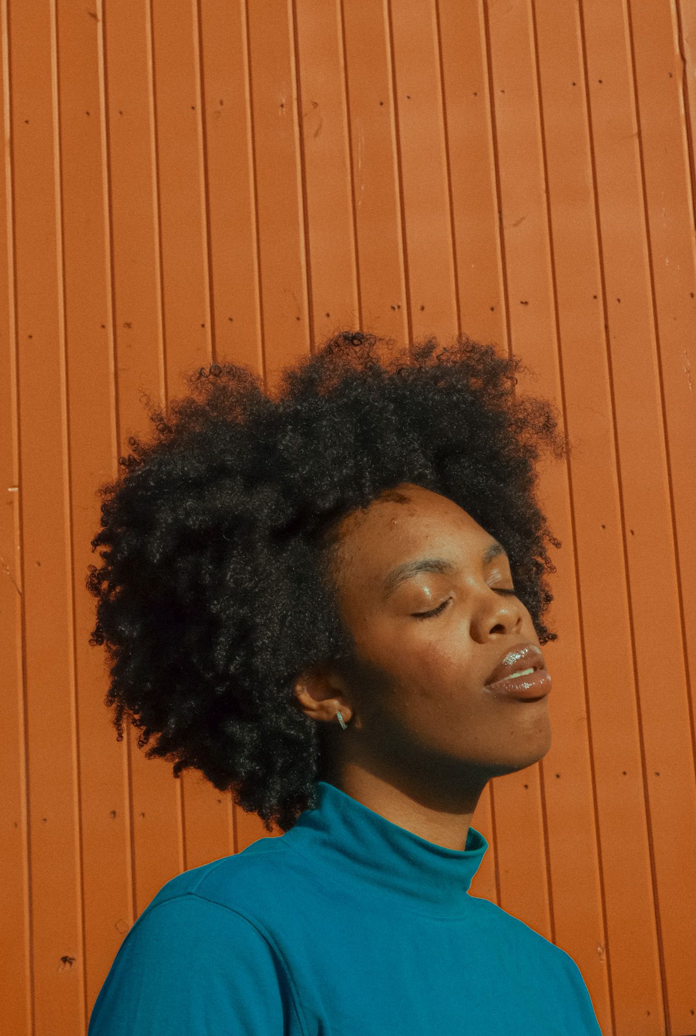 A young Black woman with natural afro hair closes her eyes in sunlight, wearing a teal turtleneck against an orange wall.