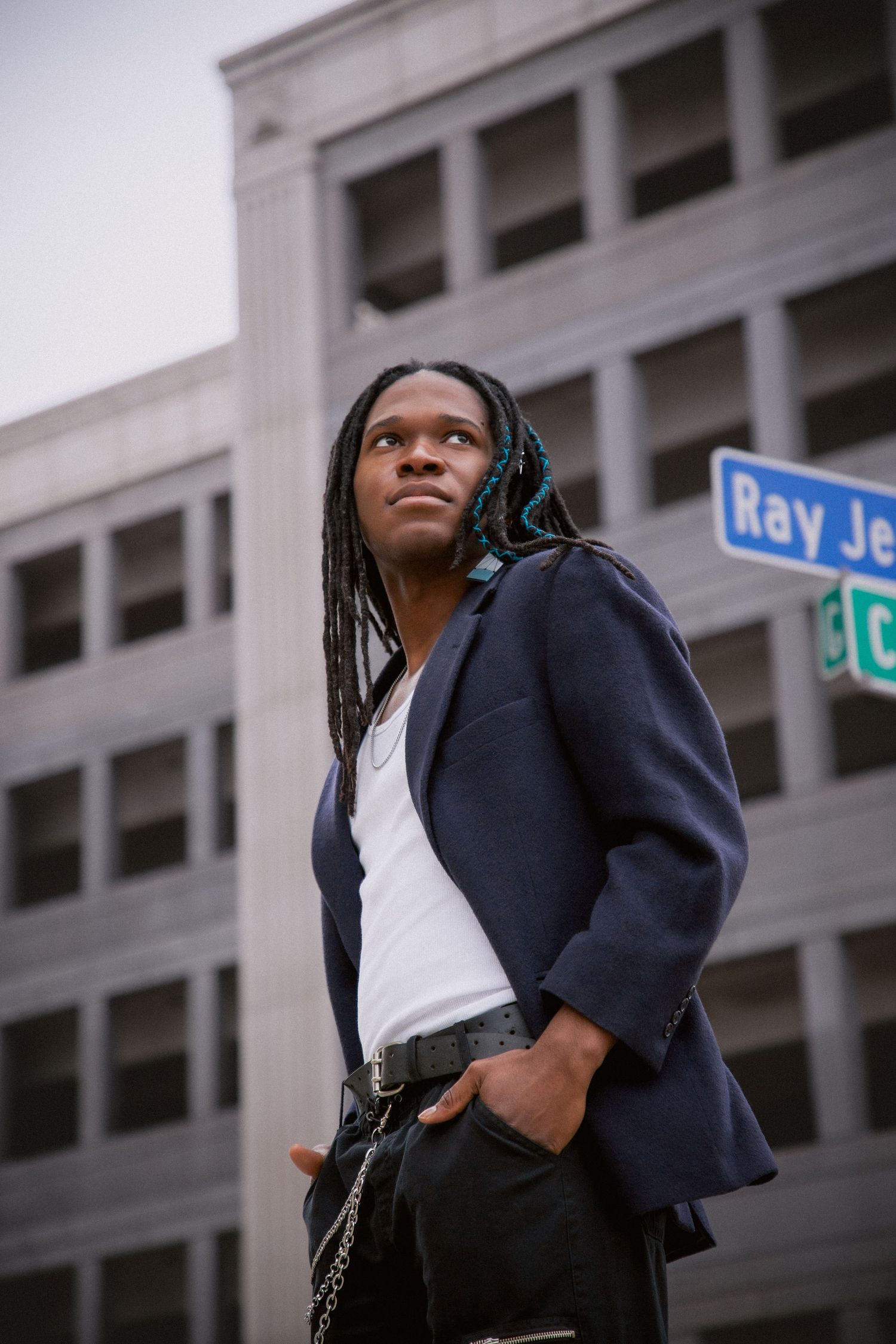 Young man with long braids in navy blazer poses confidently on urban street corner near Ray Je sign.