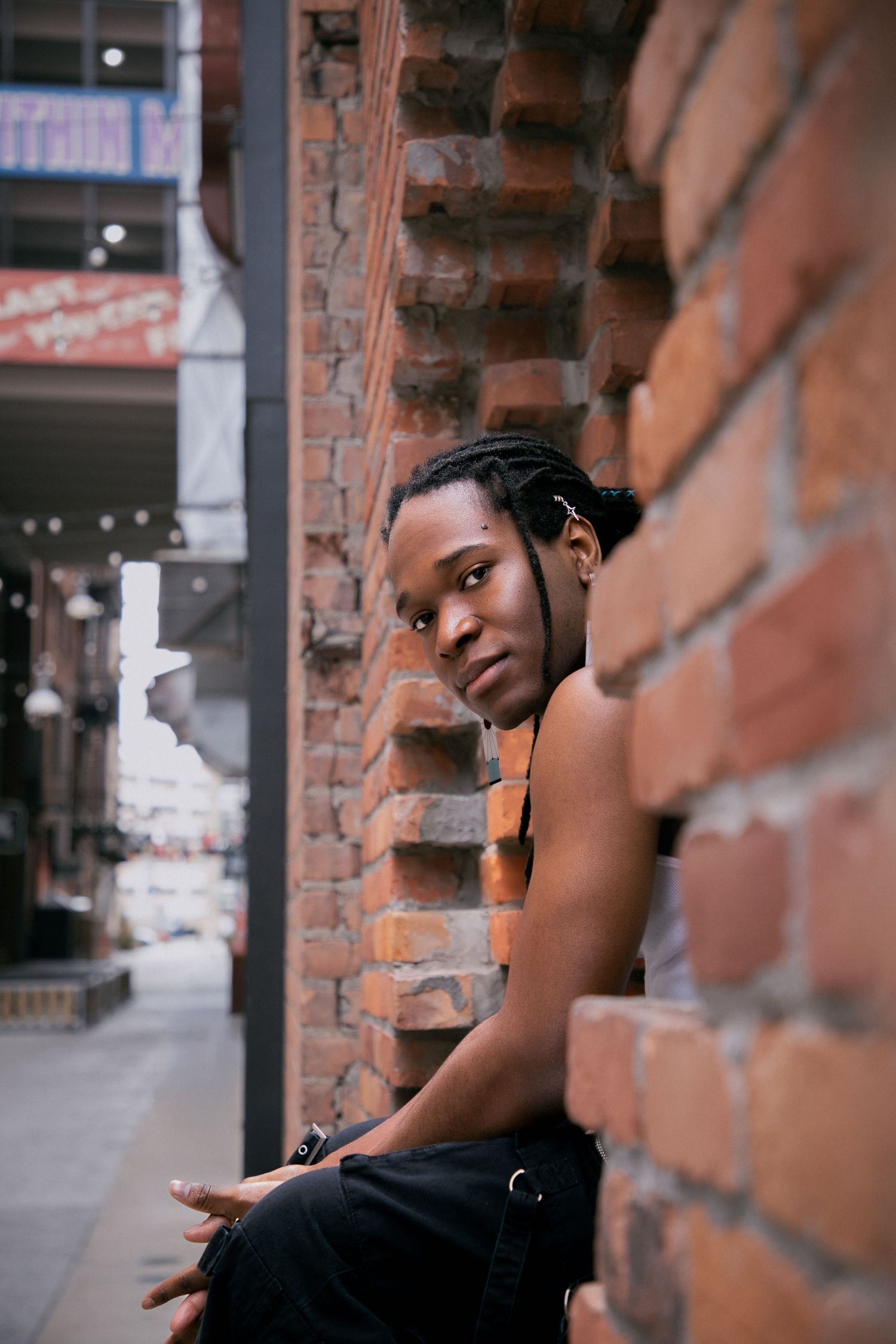 Young Black person with braids leans against a rustic brick wall in an urban alley, wearing black pants and suspenders.