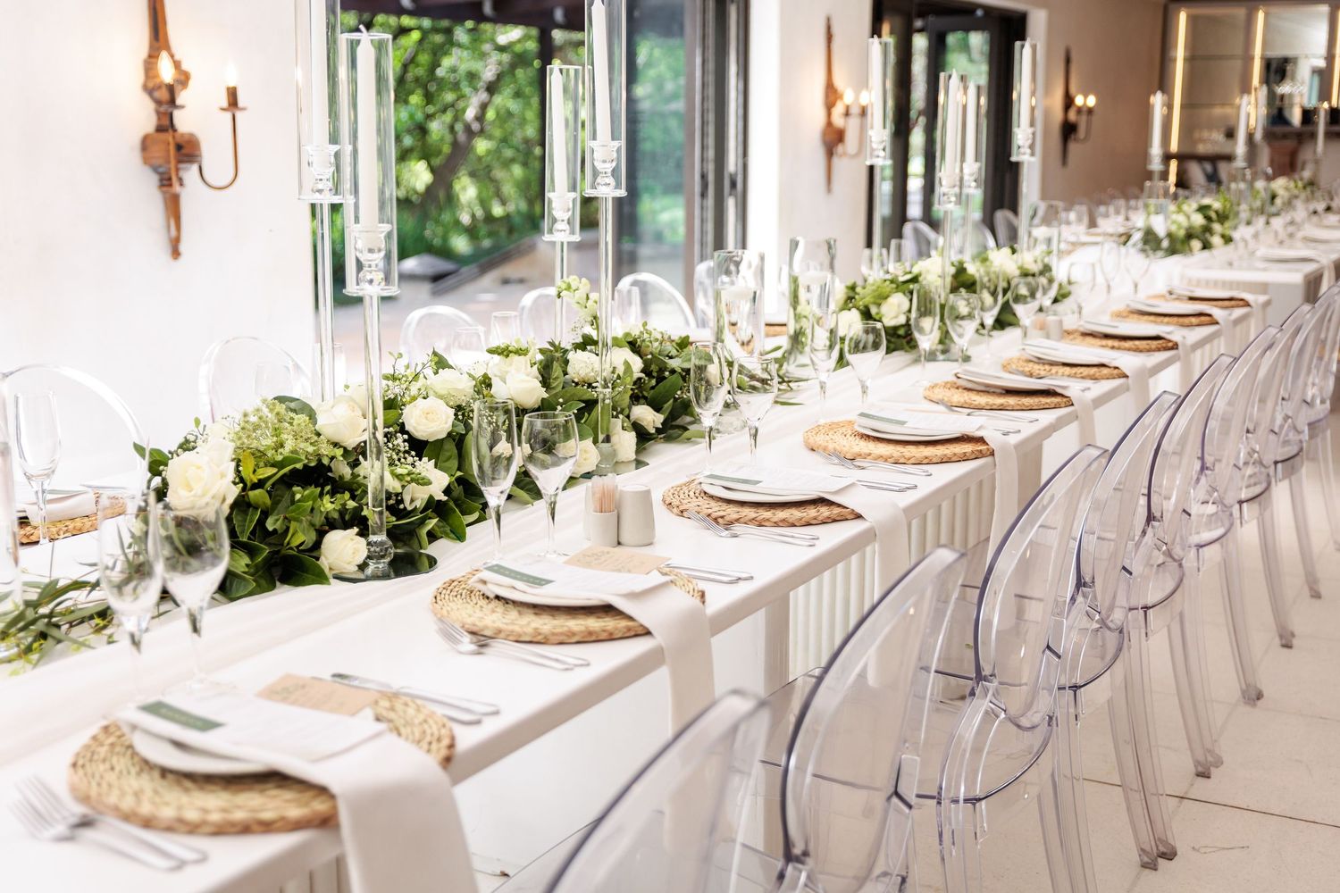 Elegant wedding reception table setup with ghost chairs, woven chargers, and white floral centerpieces in a bright venue.