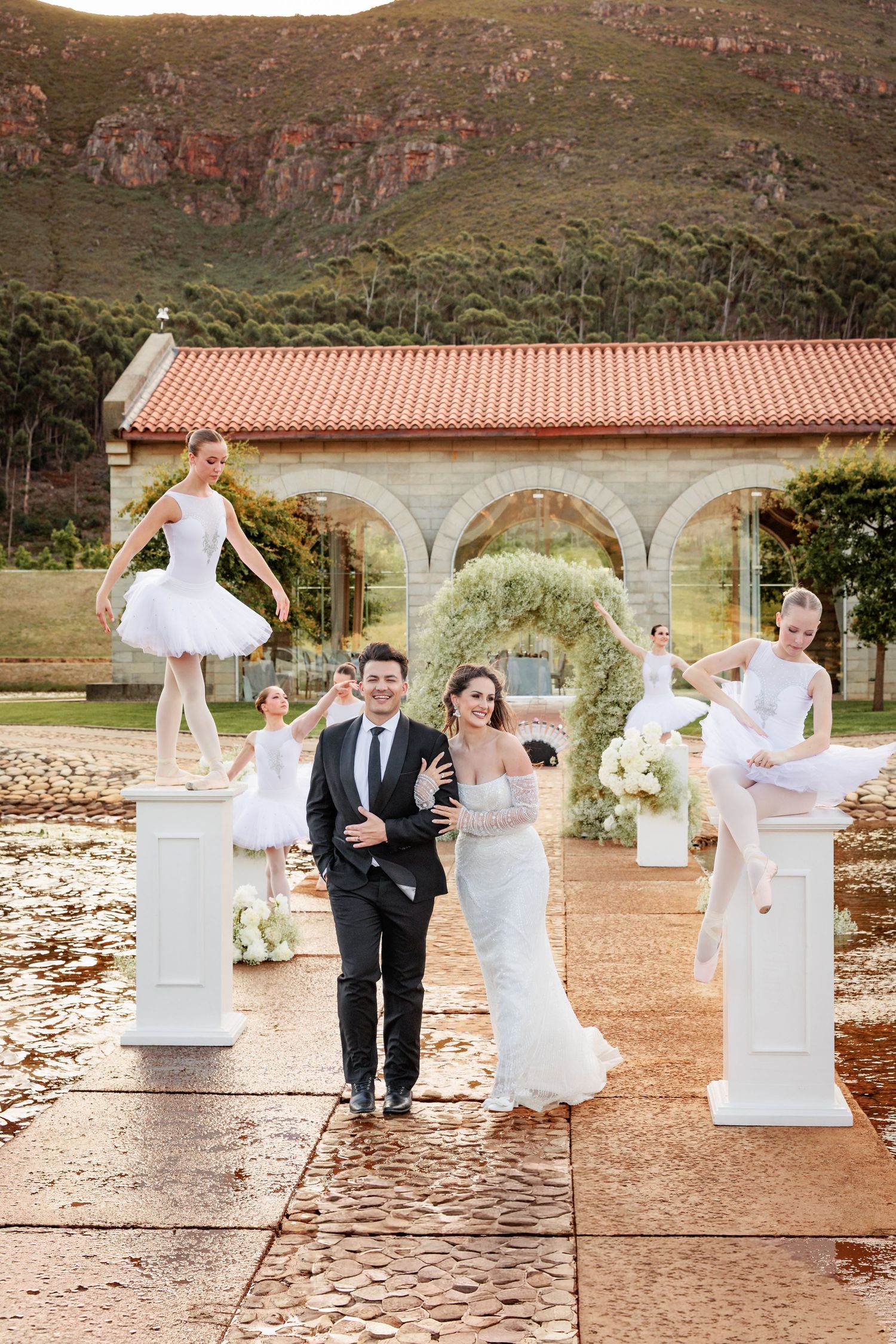 Wedding party poses playfully between white stone pillars at outdoor venue with mountain backdrop and terra cotta roof.