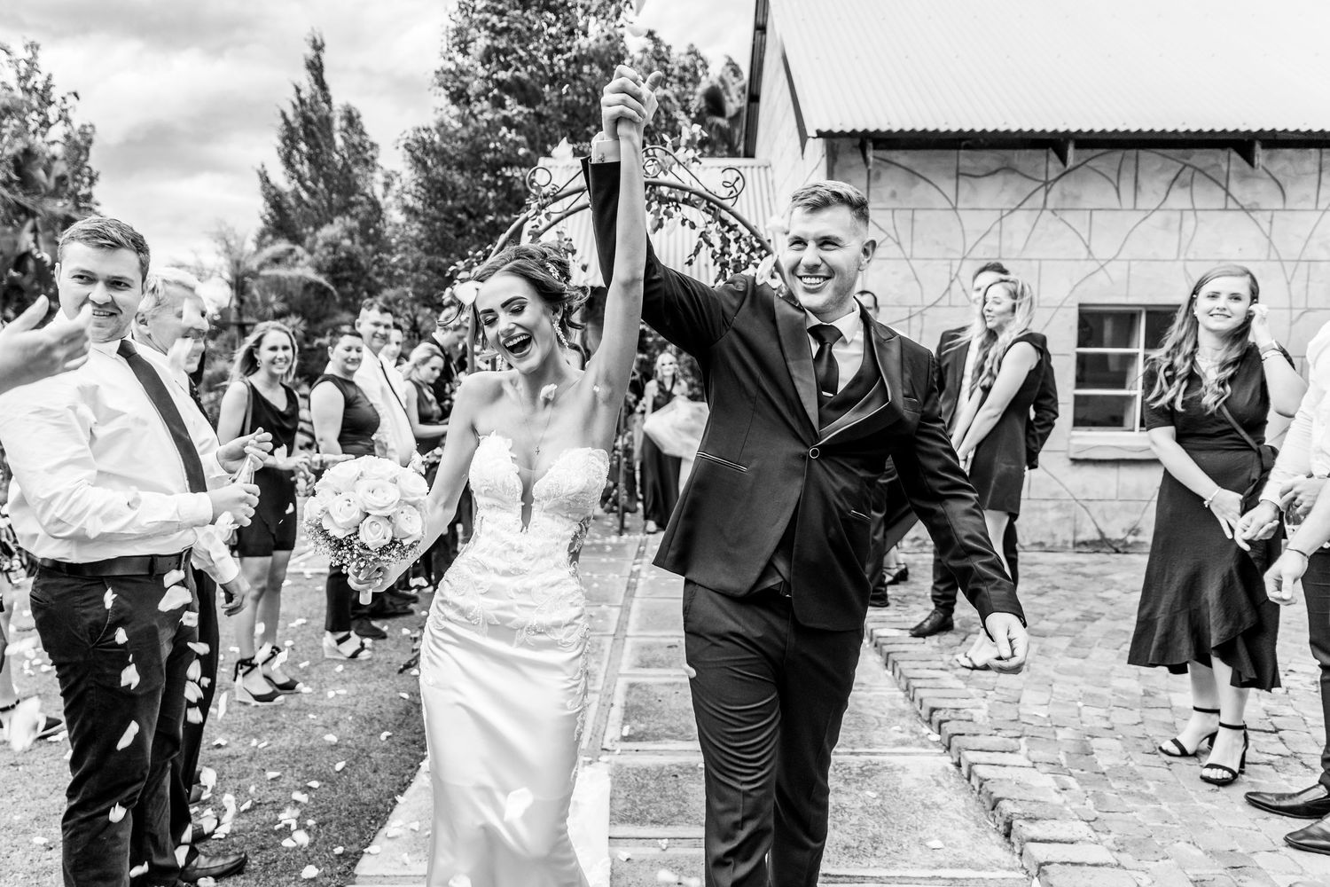 Newlyweds celebrate while walking down path lined with guests throwing confetti in black and white photo.