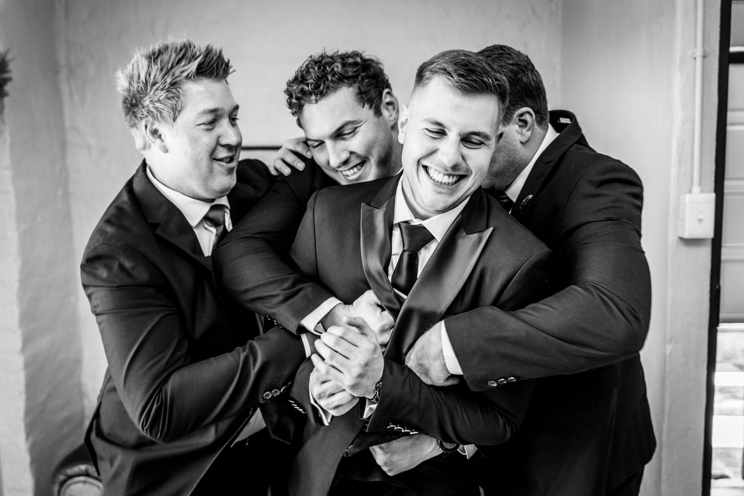 Black and white photo of three groomsmen in suits laughing and embracing at a wedding celebration.