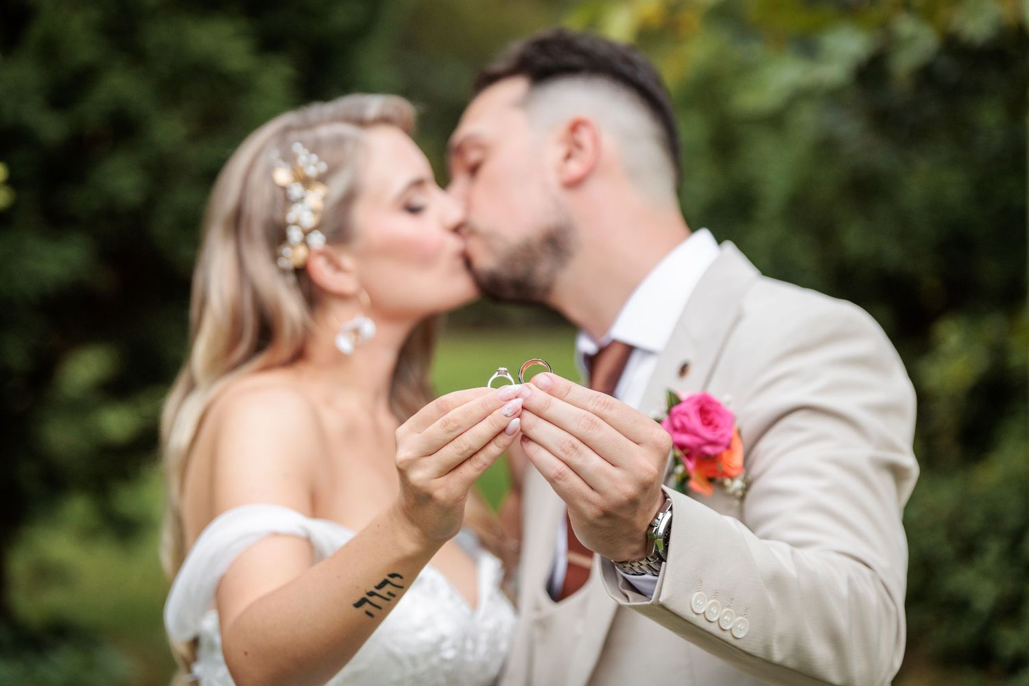 Newlyweds kissing while holding up wedding rings, bride in white dress with floral hair accessories, groom in beige suit.