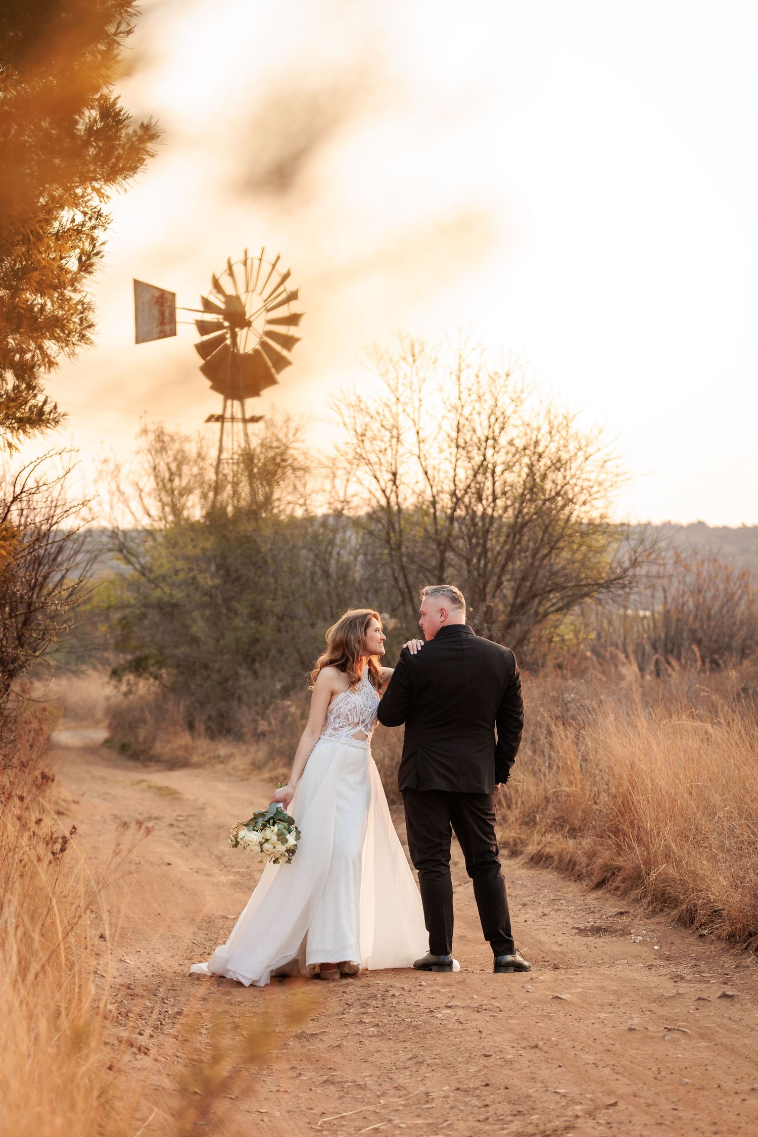 Couple shares romantic moments at sunset with rustic windmill silhouette in background on desert path.