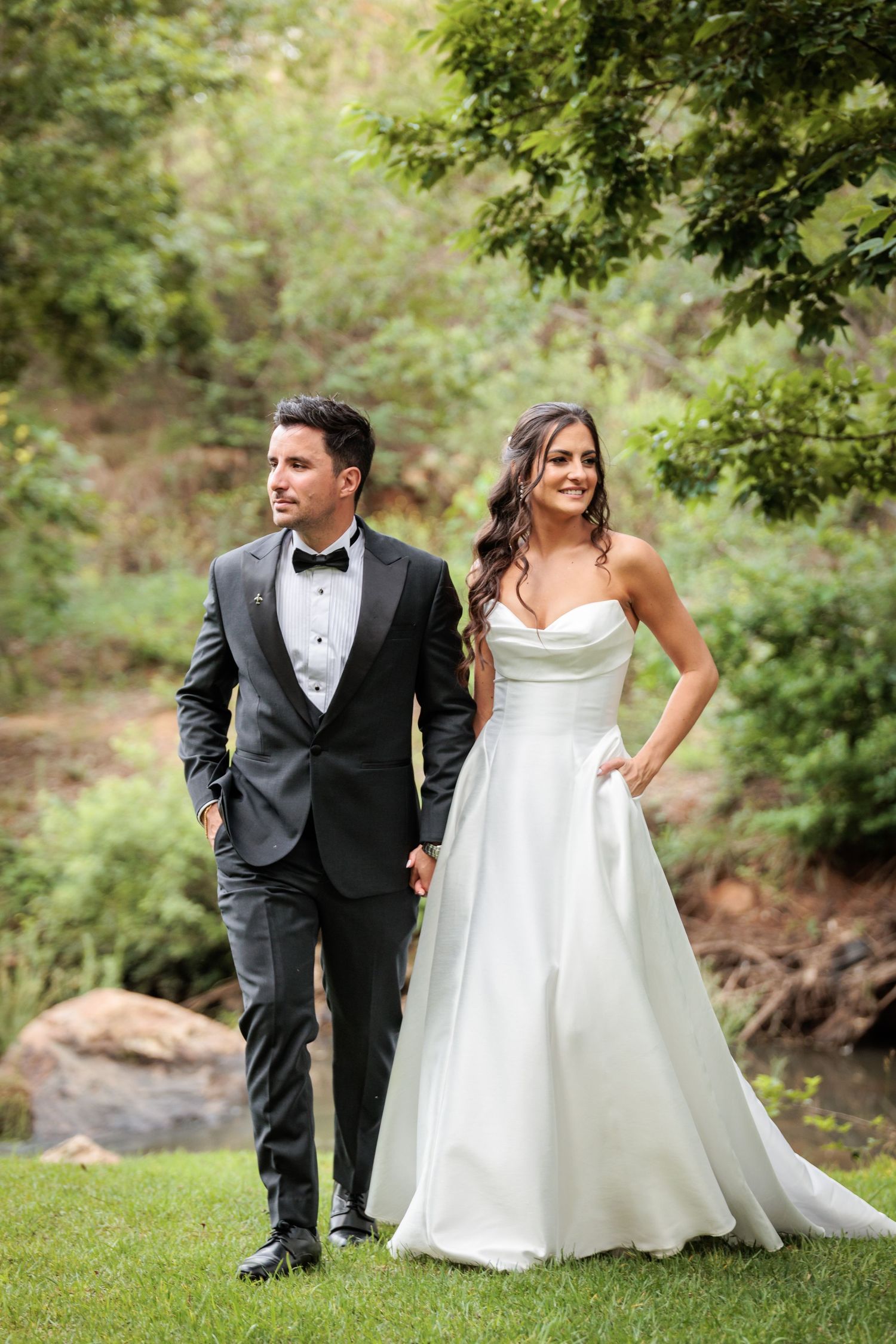 A couple in formal wedding attire stands together on grass with lush greenery in the background.