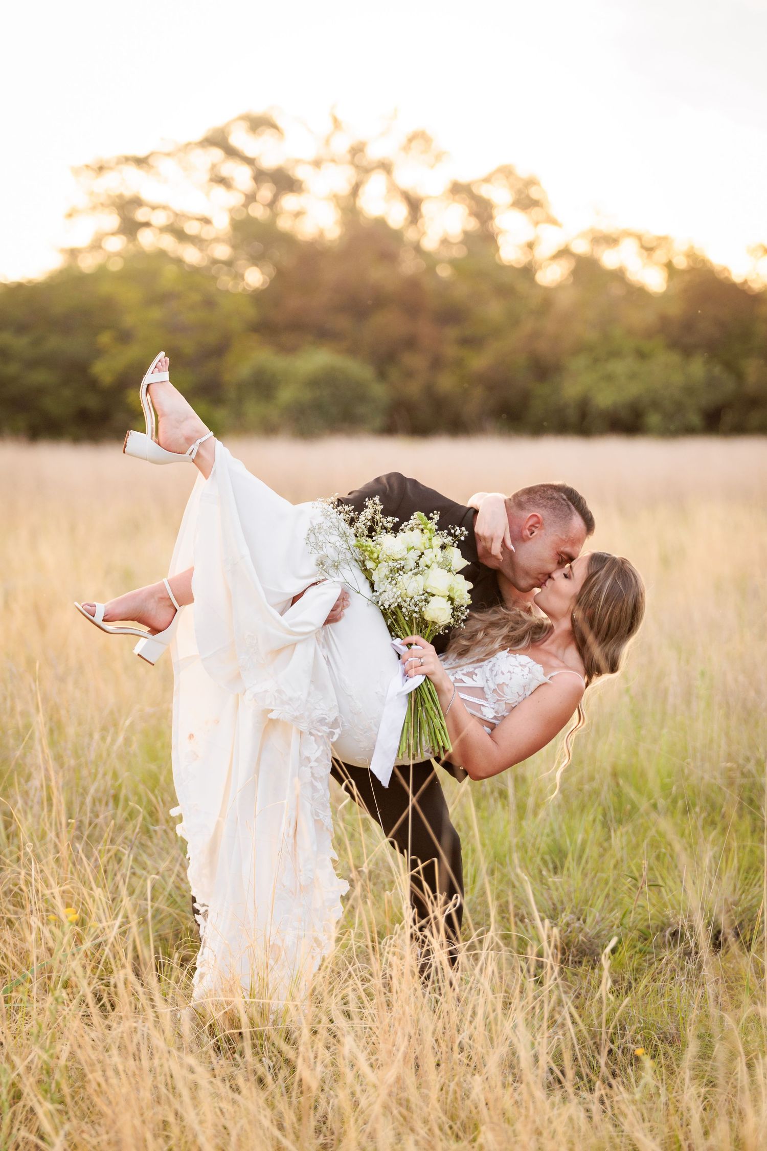 A romantic wedding couple embraces in a golden sunlit field during sunset while holding a white floral bouquet.