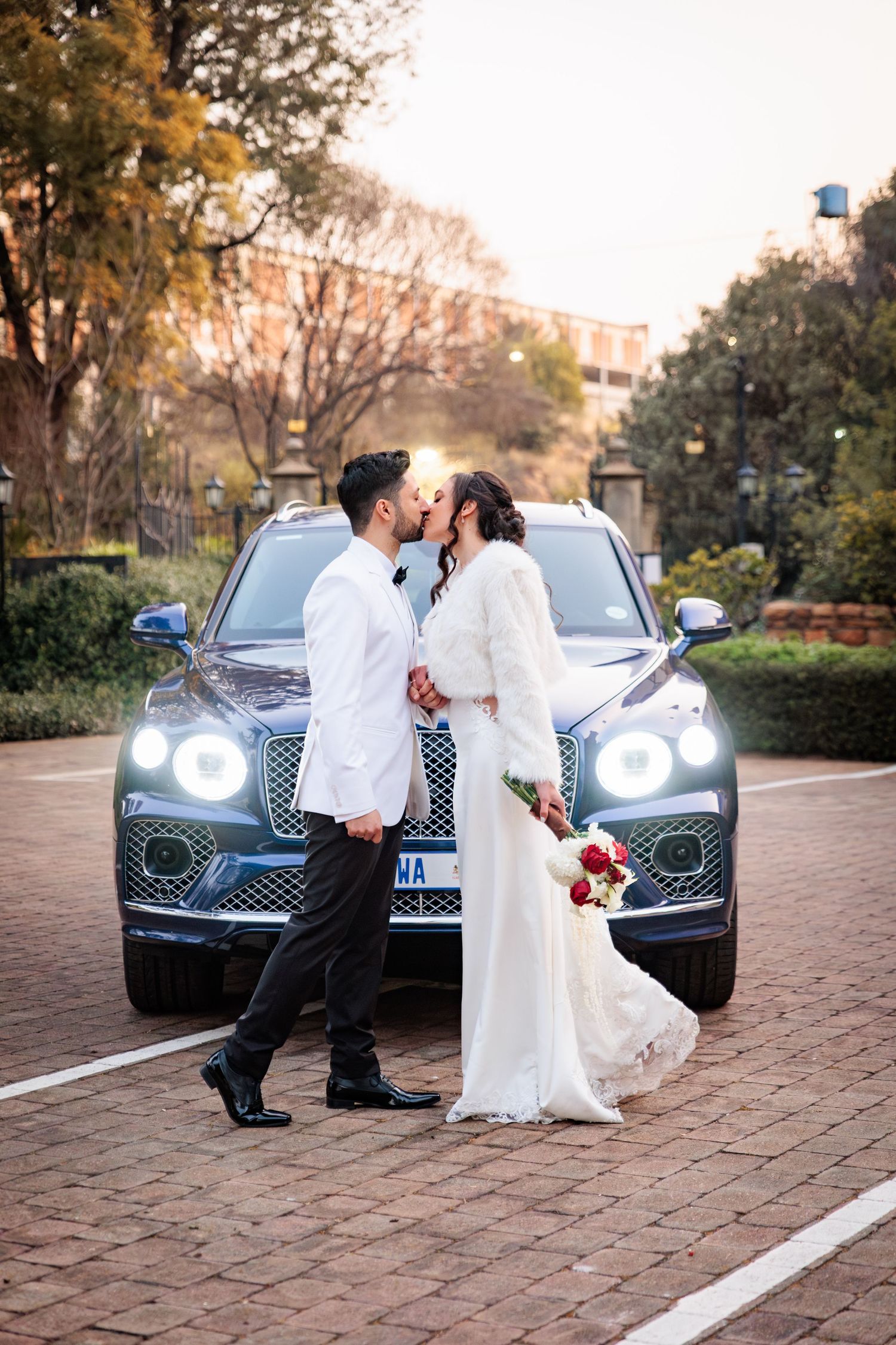 A romantic moment shared between a couple in front of a luxury car with illuminated headlights at dusk.