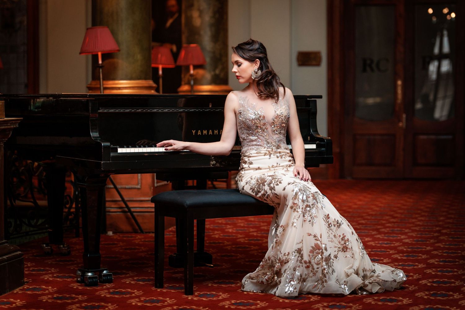 A woman in a floral dress poses gracefully near a grand piano in a dimly lit concert hall.