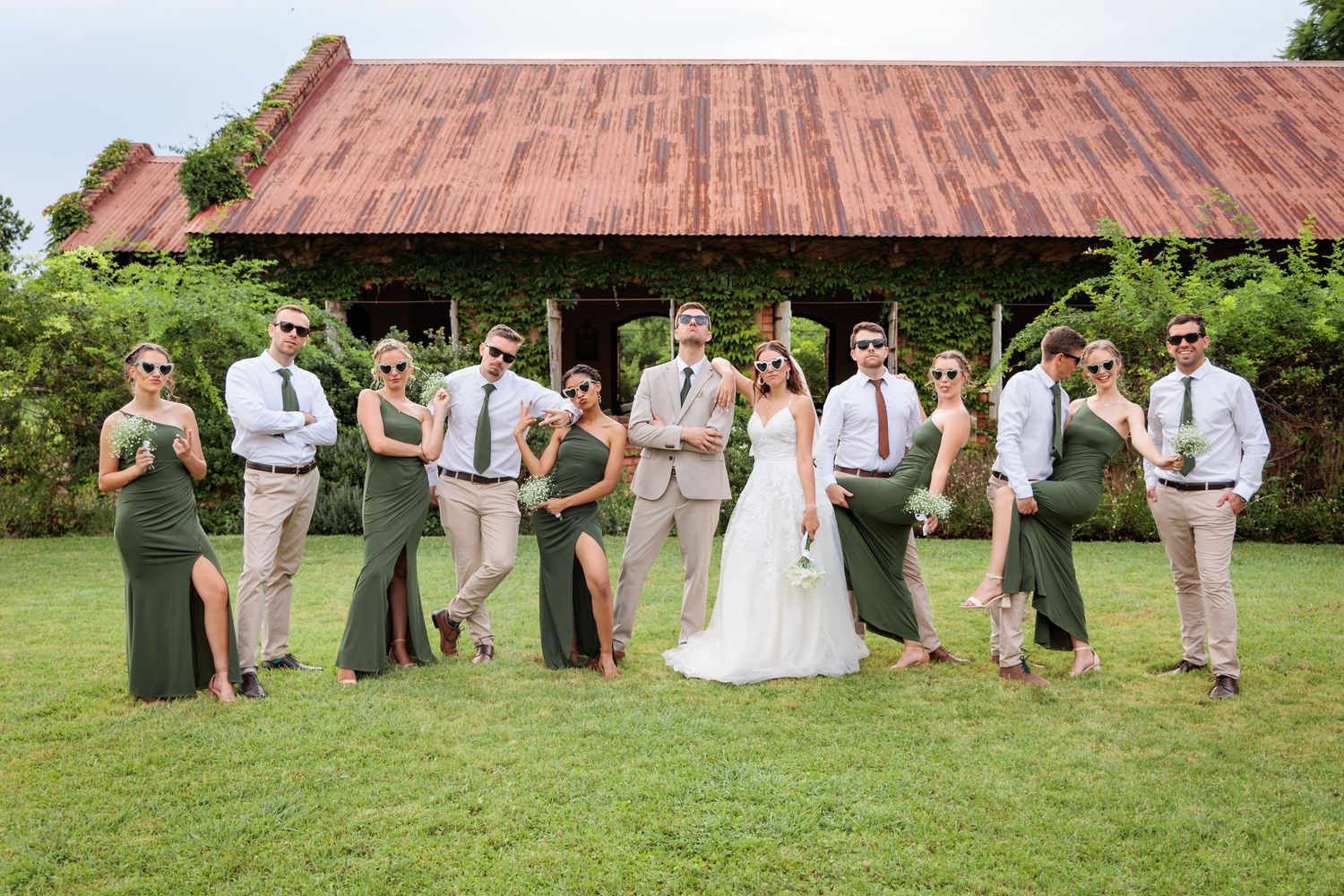 Wedding party in green dresses and tan suits posing outdoors near a rustic barn on a sunny day.