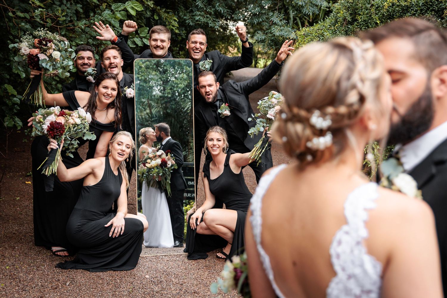 Wedding party holds a mirror reflecting the bride and groom kissing, while bridesmaids and groomsmen celebrate outdoors.