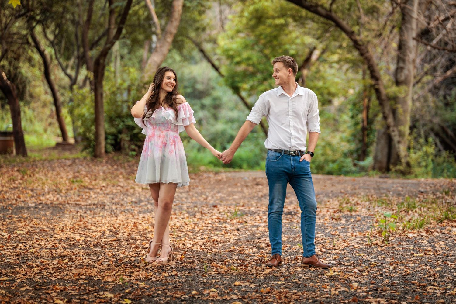 Two people walk hand in hand through a scenic autumn park covered in fallen leaves.