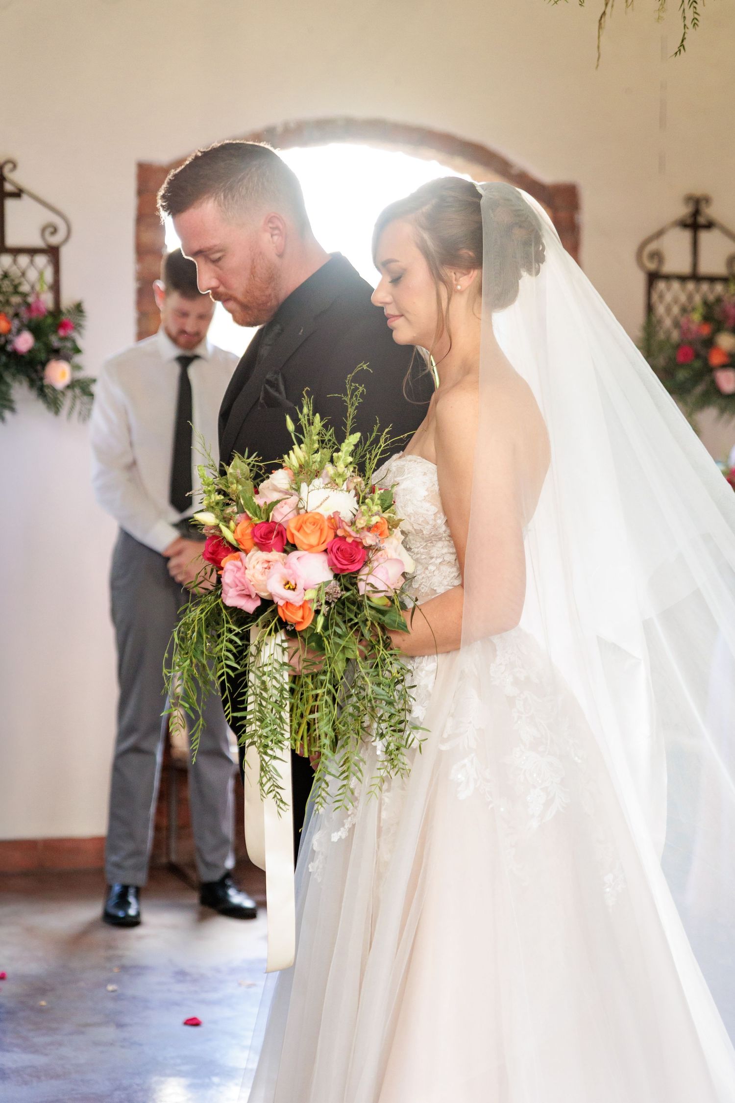 A couple stands at the altar during their wedding ceremony, with the bride holding a lush bouquet of pink flowers and greenery.