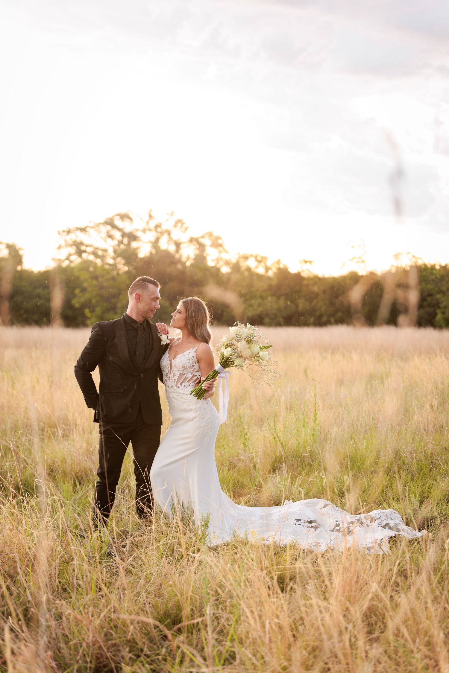 Romantic wedding couple walks through golden field at sunset during outdoor photo session.