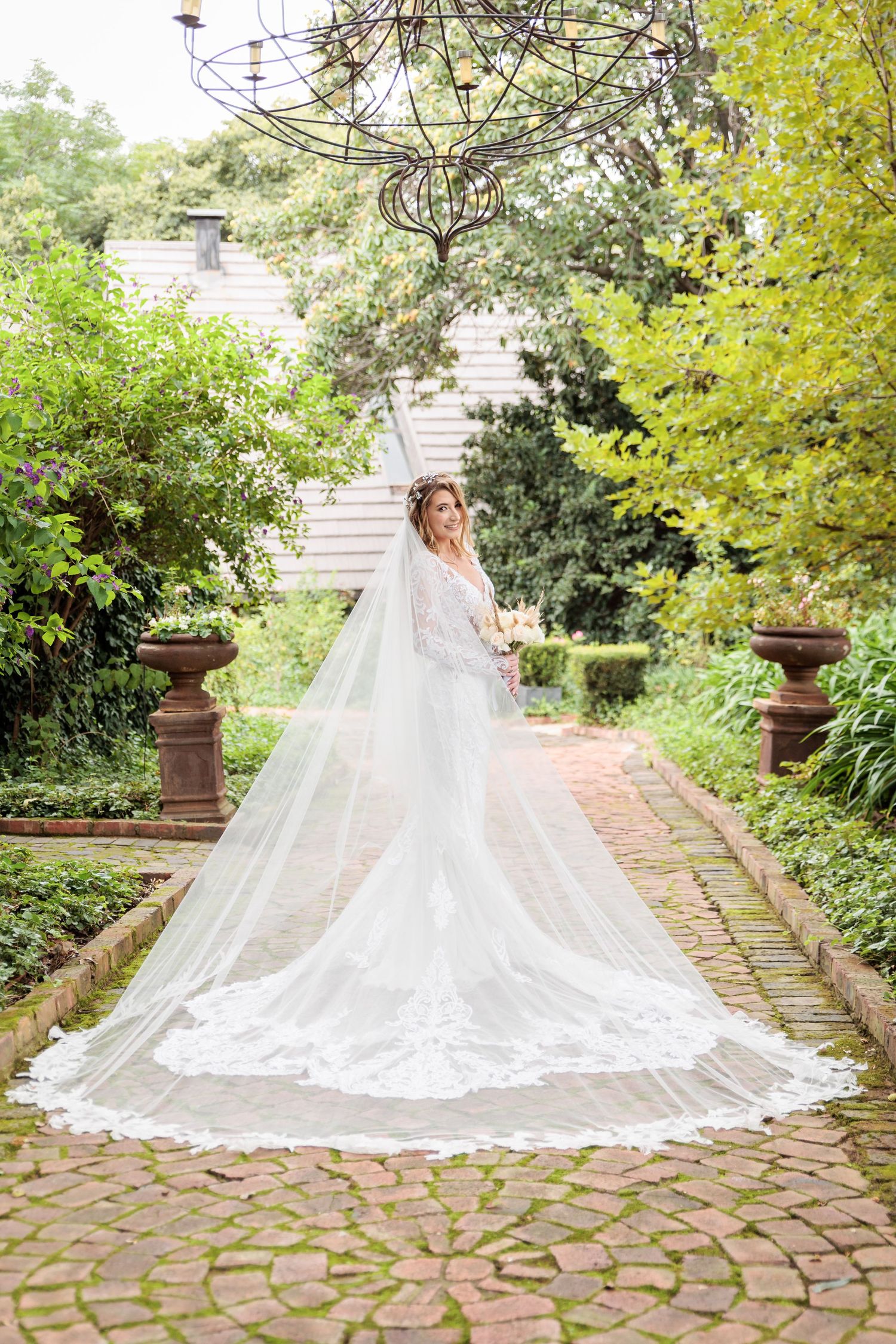 A bride in a flowing white gown and cathedral veil poses on stone steps surrounded by lush greenery in a garden setting.