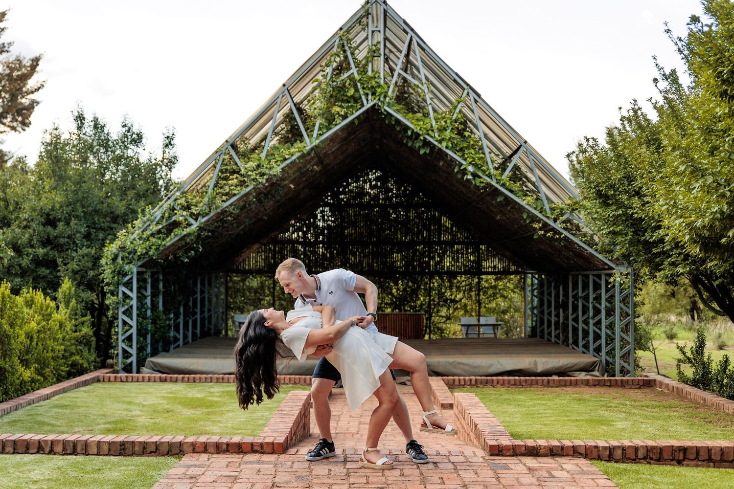 Two people playfully run and dance together on a brick walkway in an outdoor garden setting.