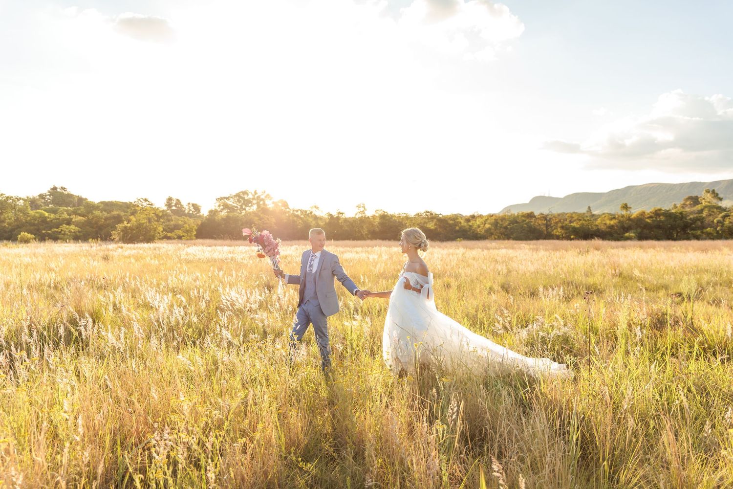 Couple walks through golden wheat field at sunset holding hands while bride's dress and veil flow in the wind.