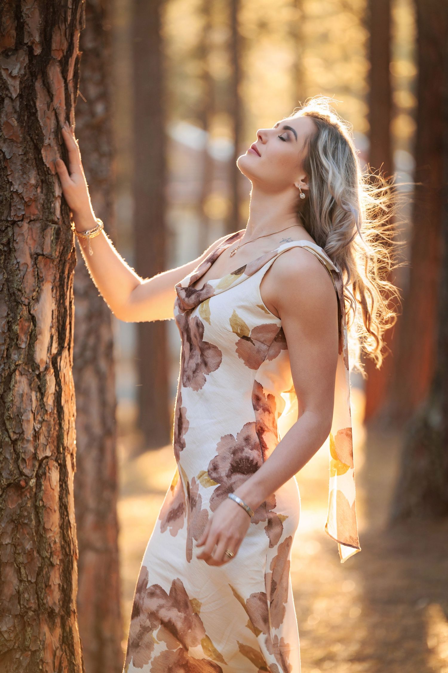 A woman in a floral dress poses gracefully against tree trunks in a sunlit forest during golden hour.