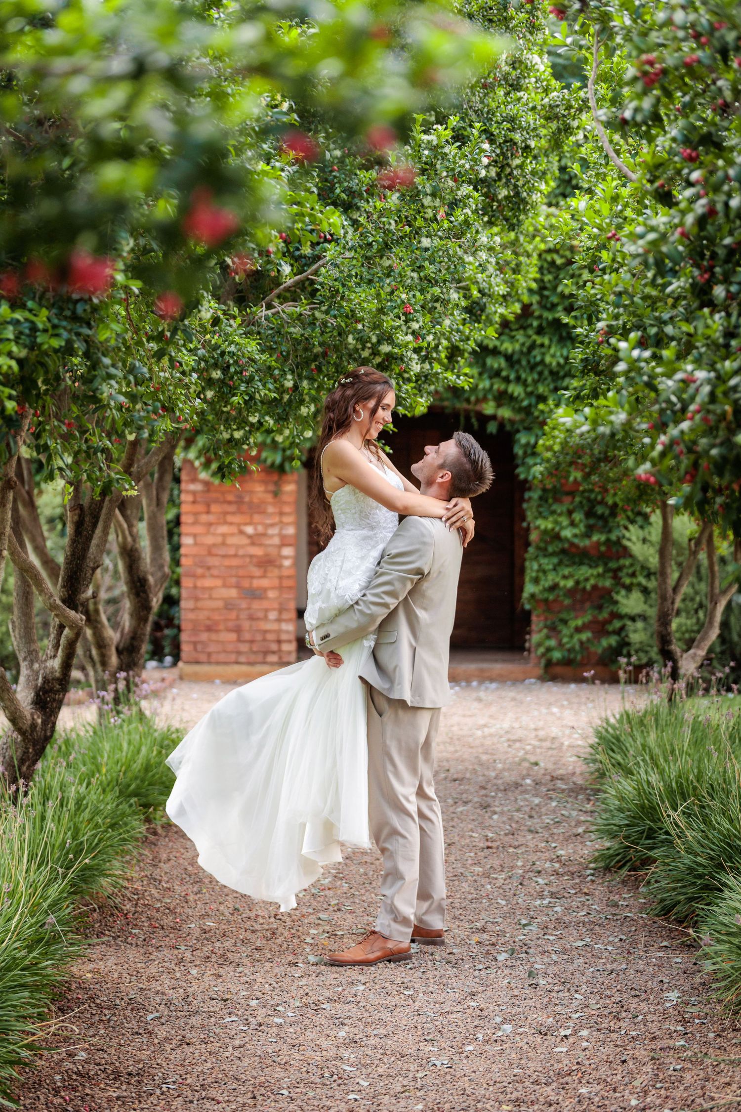 A romantic wedding couple embraces and dances in a garden path lined with green trees and red flowers.