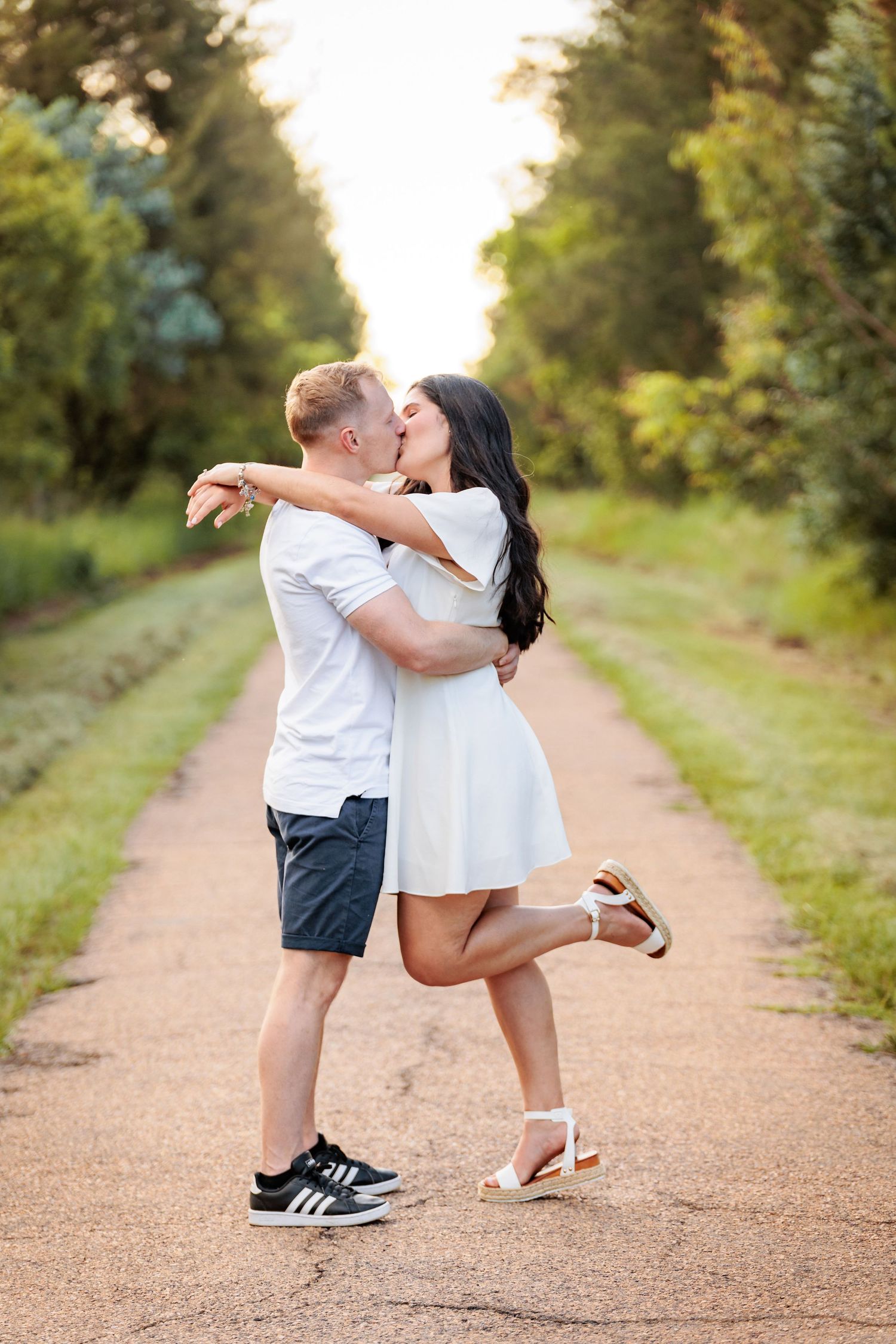 A couple shares an intimate kiss while dancing together on a gravel path surrounded by lush greenery.