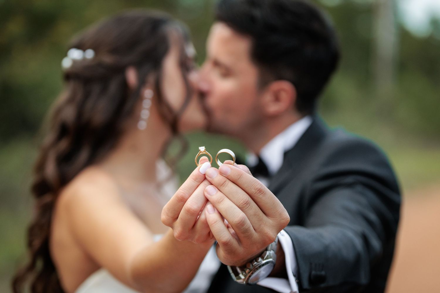 Bride and groom kissing while holding wedding rings together outdoors in a romantic blurred background portrait.
