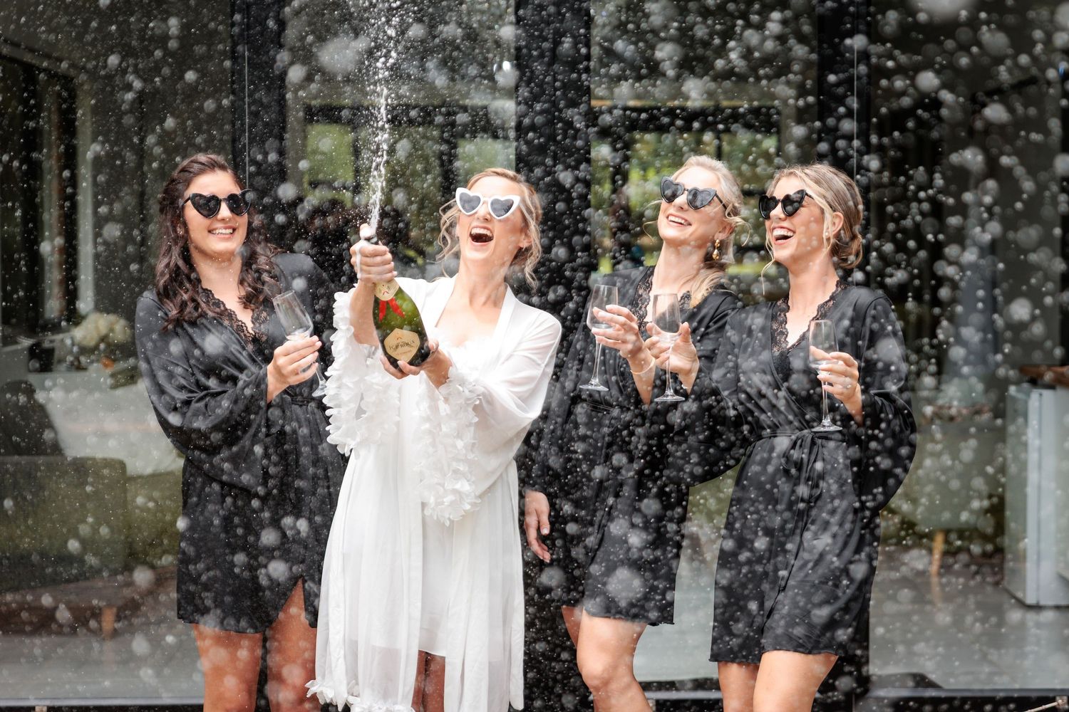 Bride in white robe pops champagne with bridesmaids in black robes, all wearing sunglasses, celebrating outdoors.