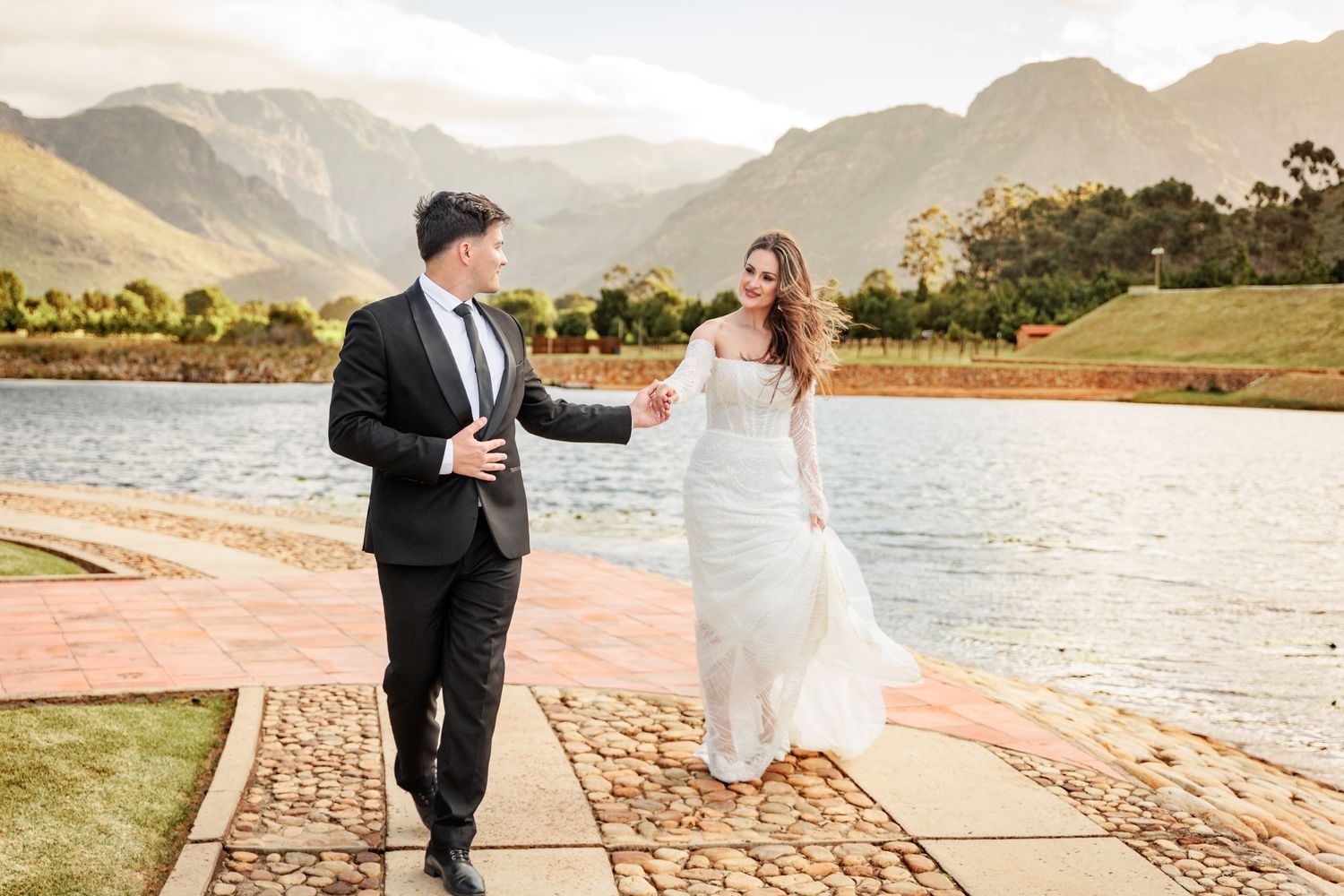 A couple in formal wedding attire walks together on a scenic lakeside path during a sunset celebration.